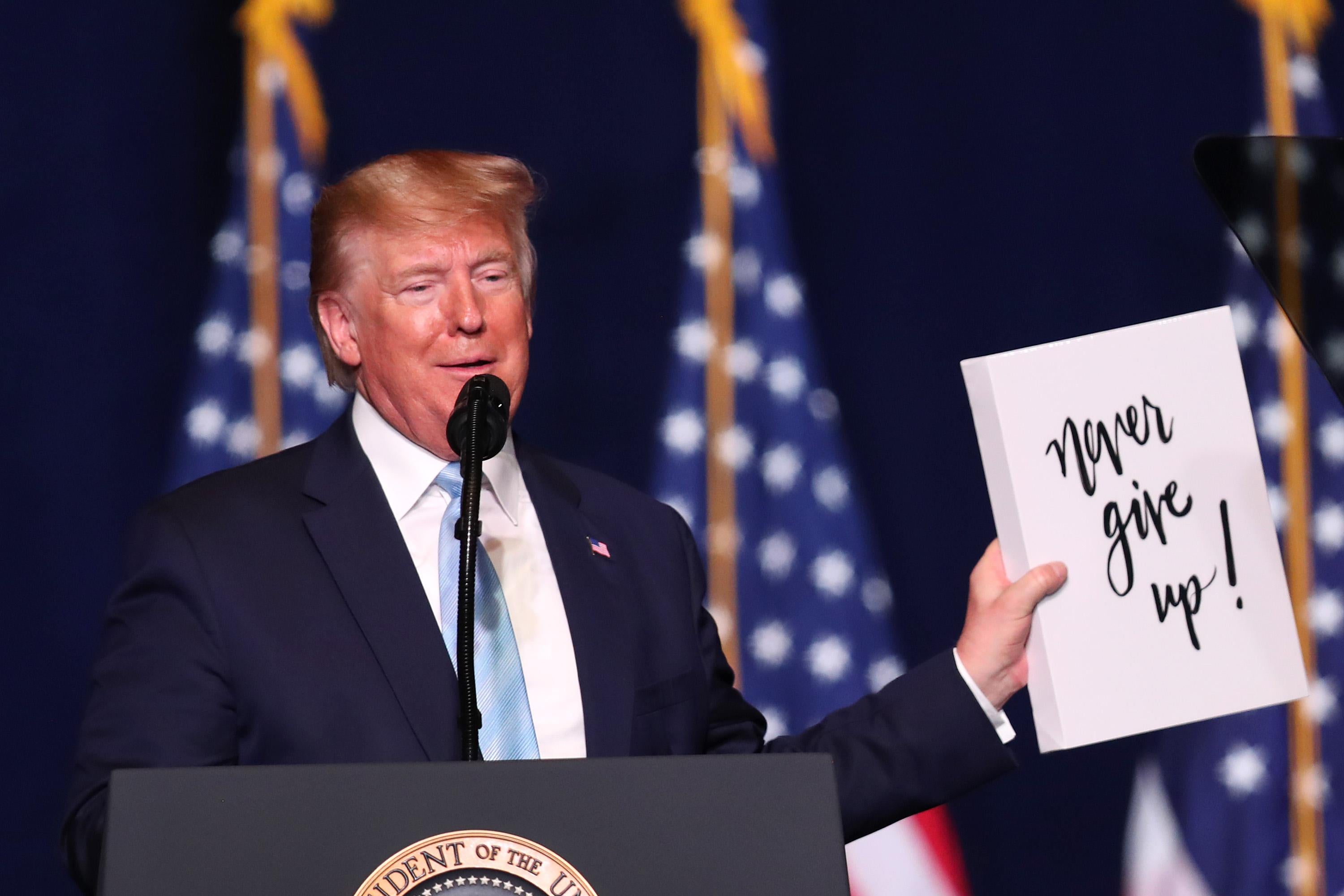 Donald Trump stands at a podium and holds up a sign that reads "Never Give Up!" in a loopy typeface