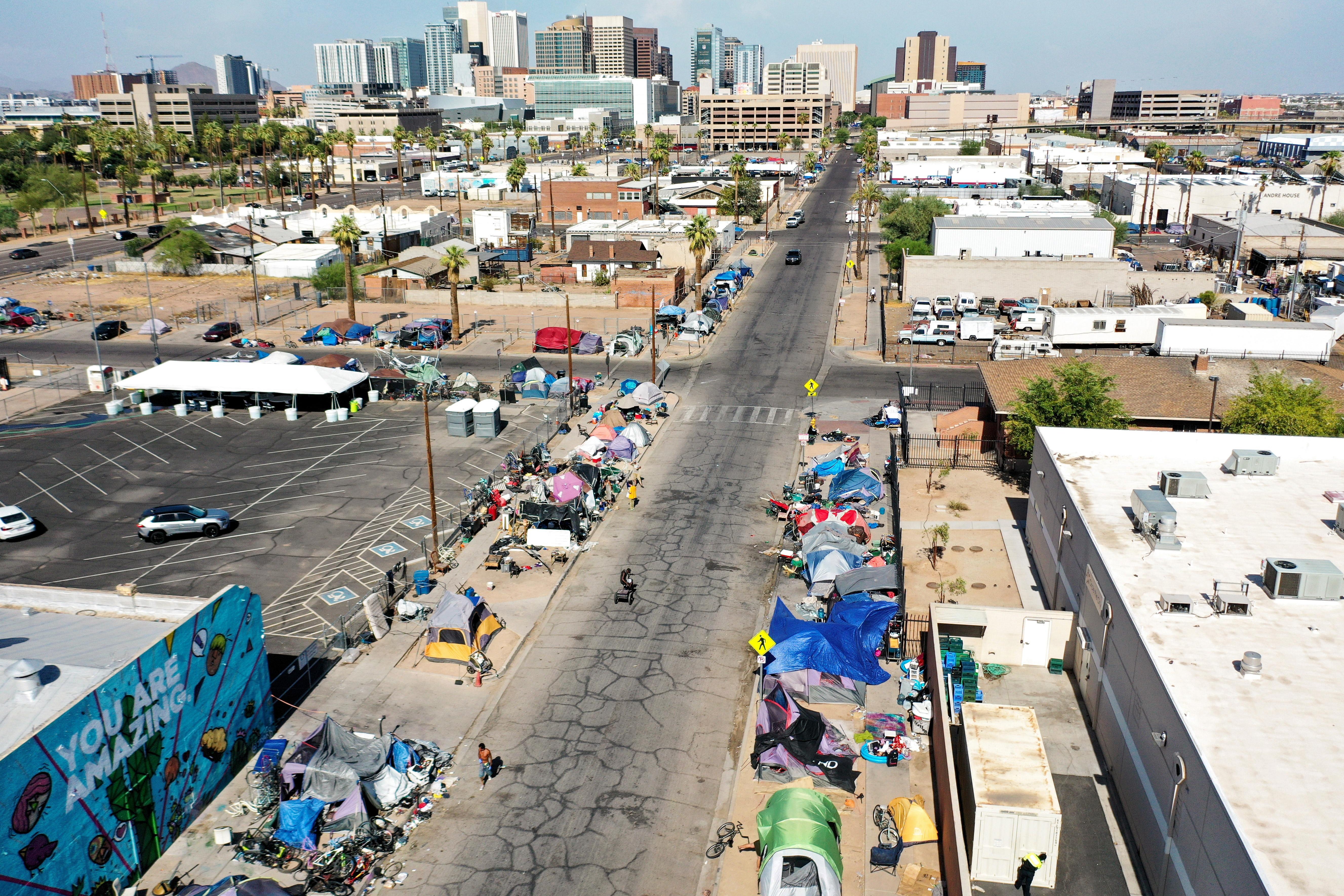 An aerial shot of Phoenix, Arizona. In the background, tall buildings from a downtown area are seen, as are palm trees. A road cuts through the middle of the photo, and on both sides are tents. 
