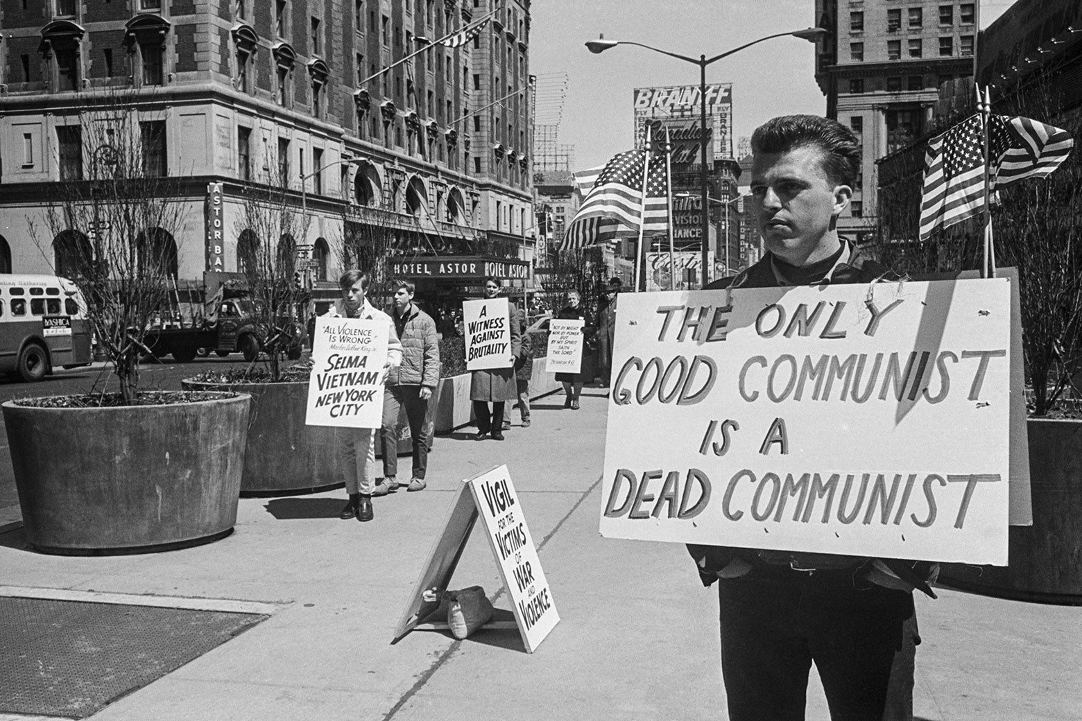 A man stands on a sidewalk wearing a sign that says "The only good Communist is a dead Communist" in front of antiwar protesters wearing signs that say "A witness against brutality" and "All violence is wrong: Selma Vietnam New York City"