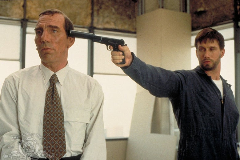 Pete Postlethwaite stands in a suit and tie looking off camera while behind him, Stephen Baldwin in a boilersuit holds a gun with a silencer to the back of his head.