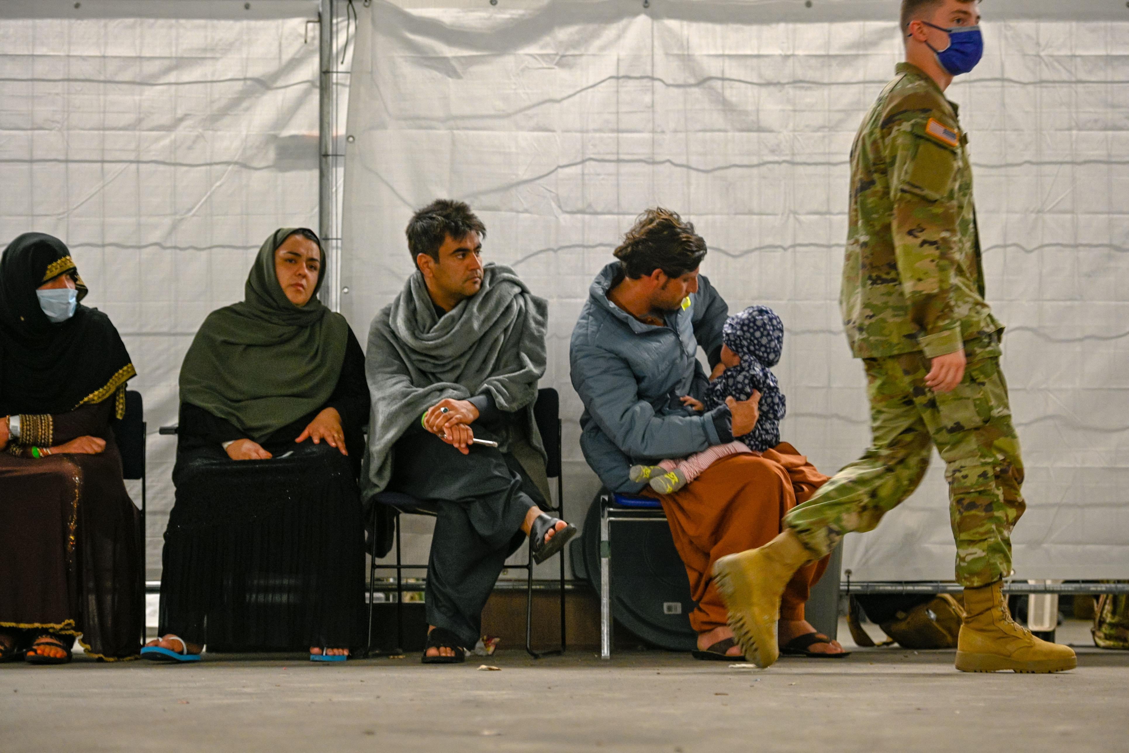 Two women in headscarves and two men, one holding a child in his lap, sit in folding chairs as a man in military fatigues and a mask walks by.