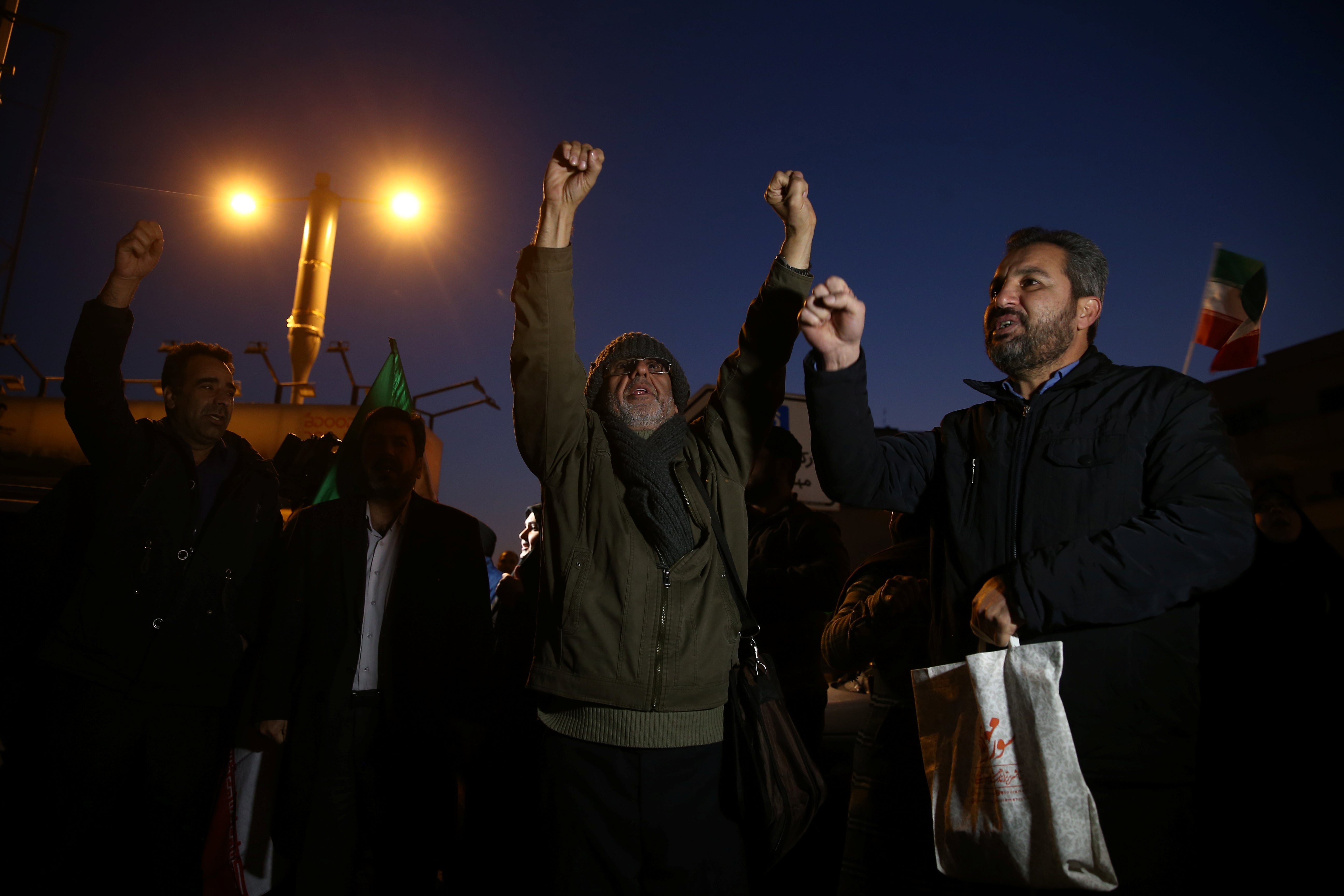 A group of men raise their fists and cheer. An Iranian flag flies in the background.