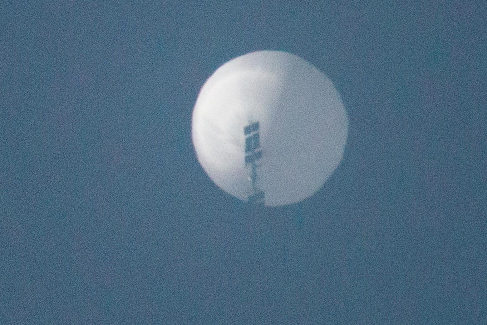 A blurry image of a large white balloon, under which black mechanical equipment is hanging, against a blue sky.