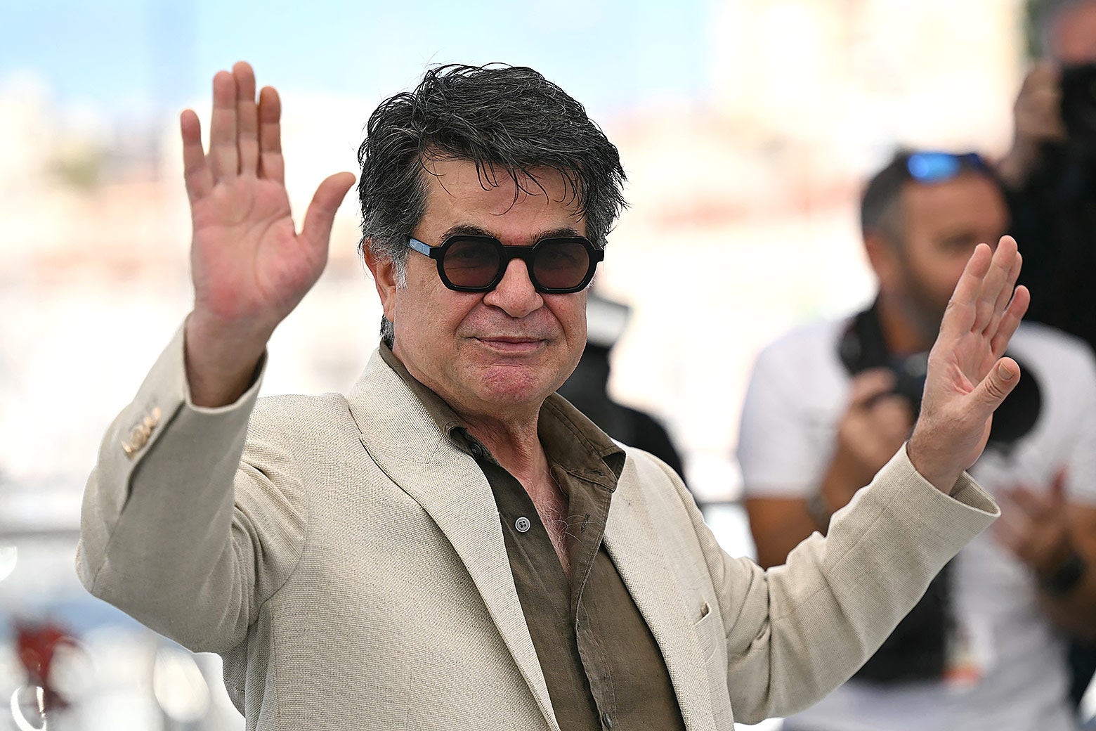 Panahi, wearing a tan suit and sunglasses, waves with both hands on the red carpet.