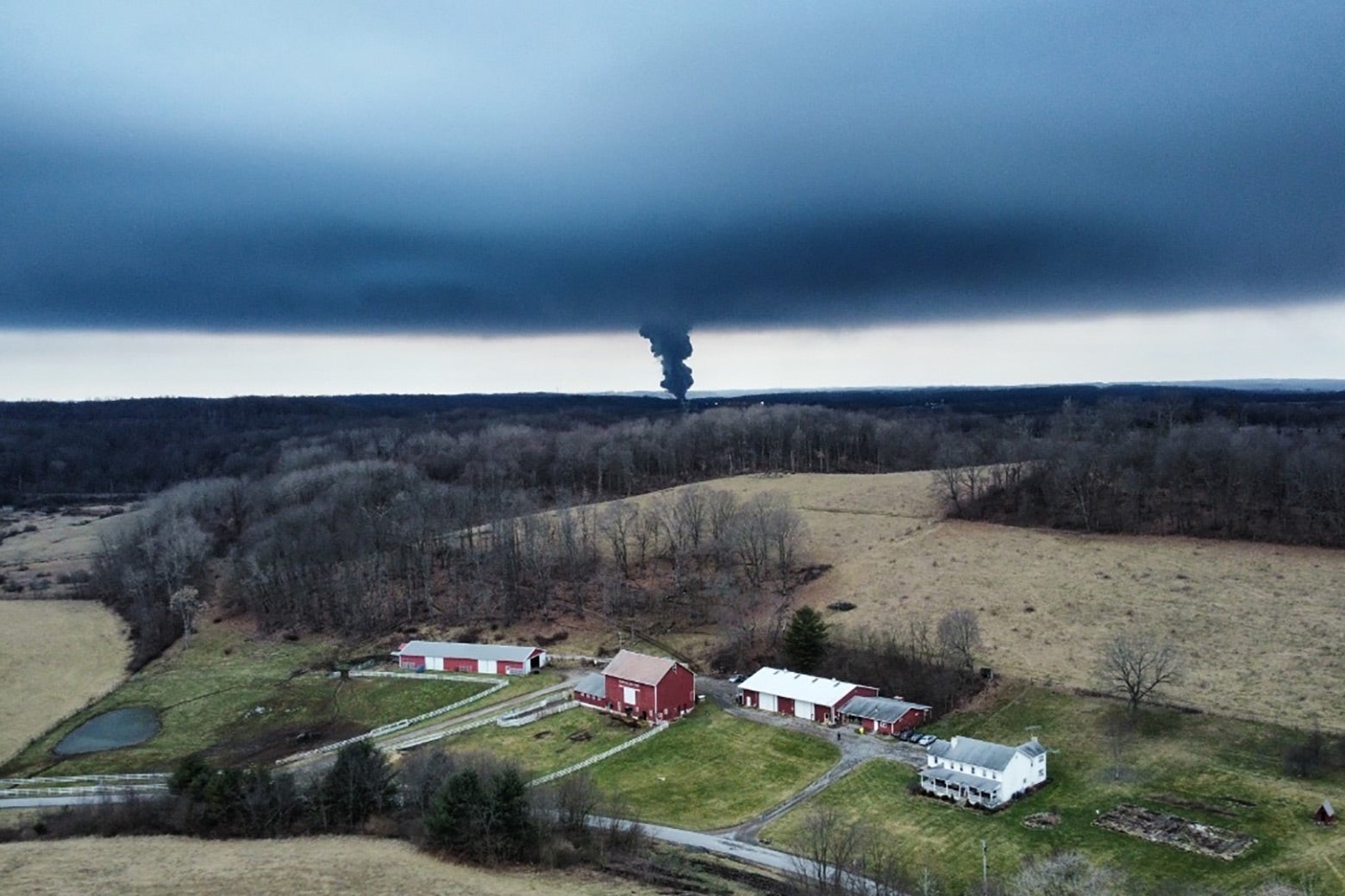 A thick plume of black smoke seen from a distance over farmland. 