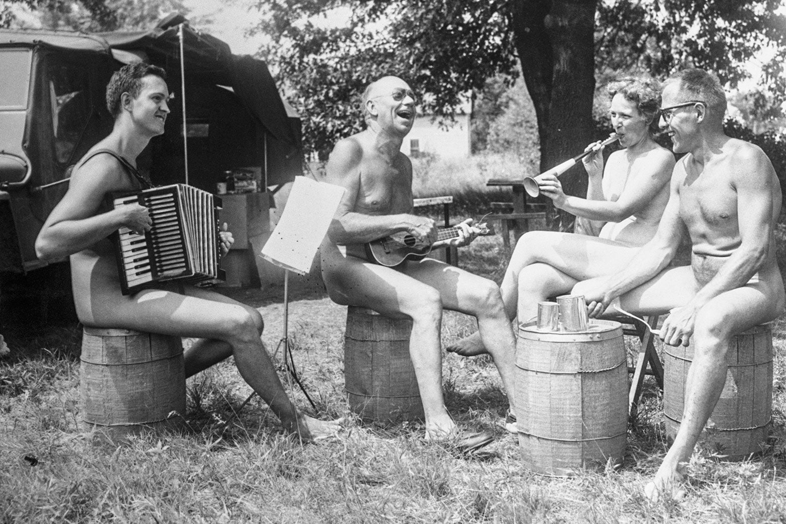 A vintage black and white photo of nudists playing instruments and sitting on barrels outdoors.