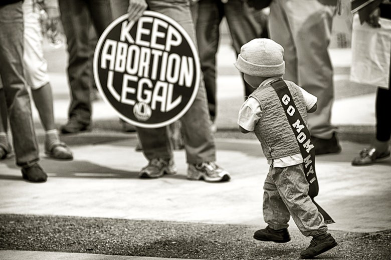 A child wearing a sash that says, "GO MOMMY," runs by someone holding a sign that says, "Keep abortion legal."