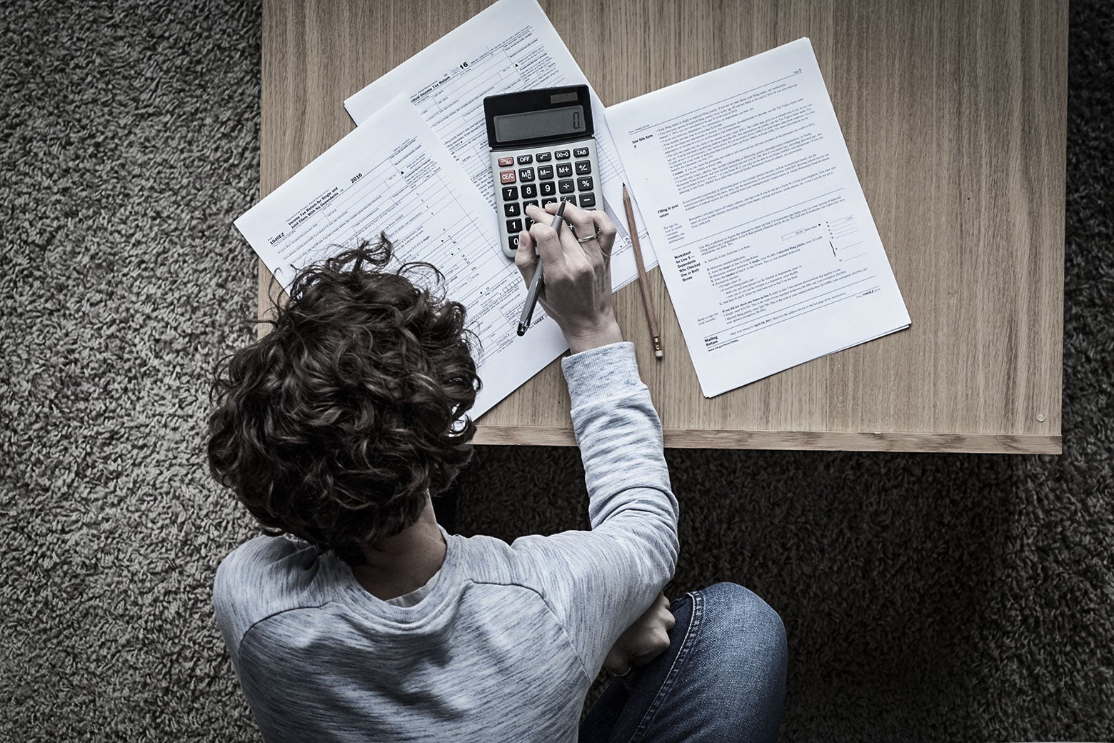 A man sits at a desk holding a pen, tax documents and a calculator spread out in front of him.