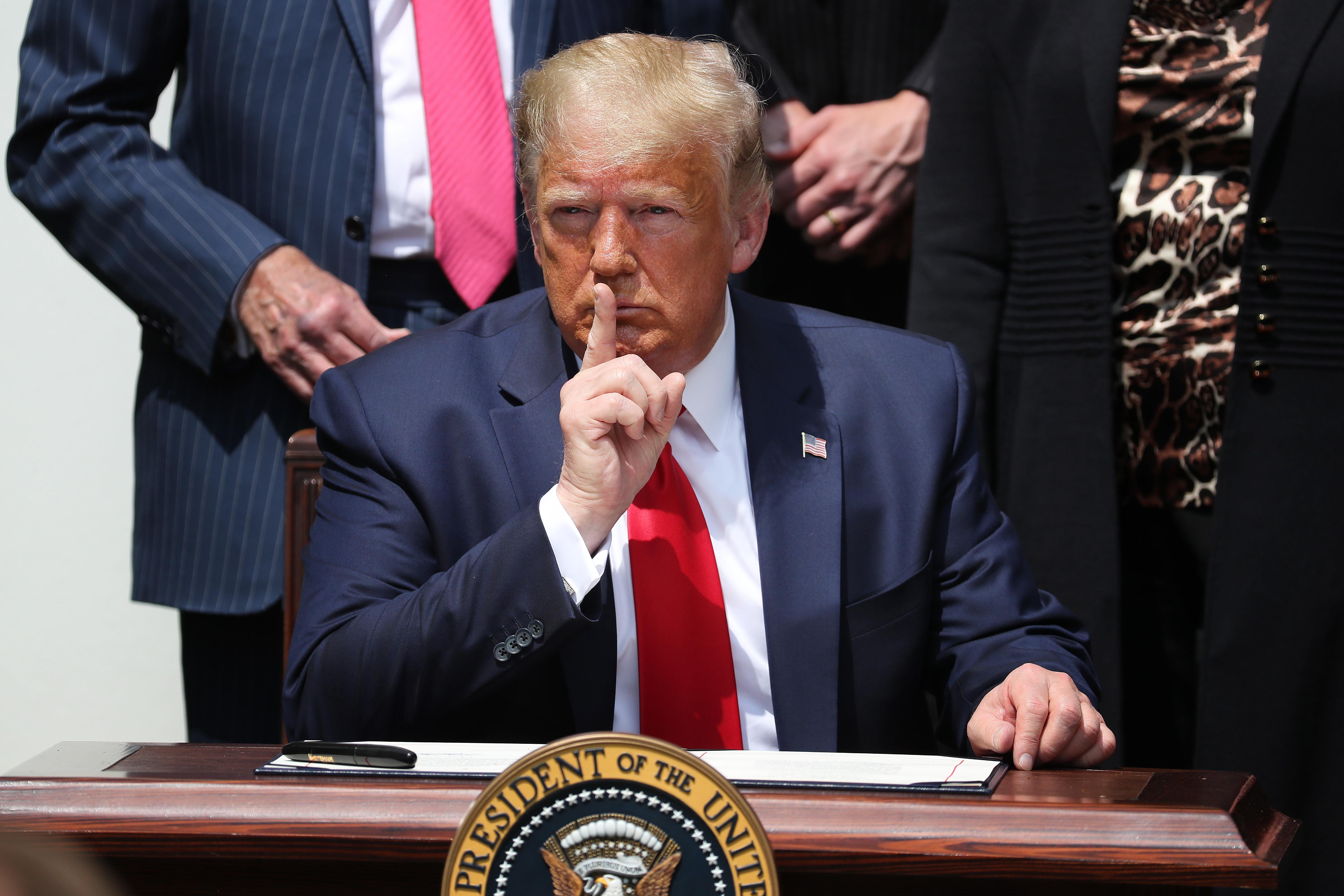 Trump holds his index finger up to his mouth in a shushing signal while sitting at a desk in the Rose Garden.