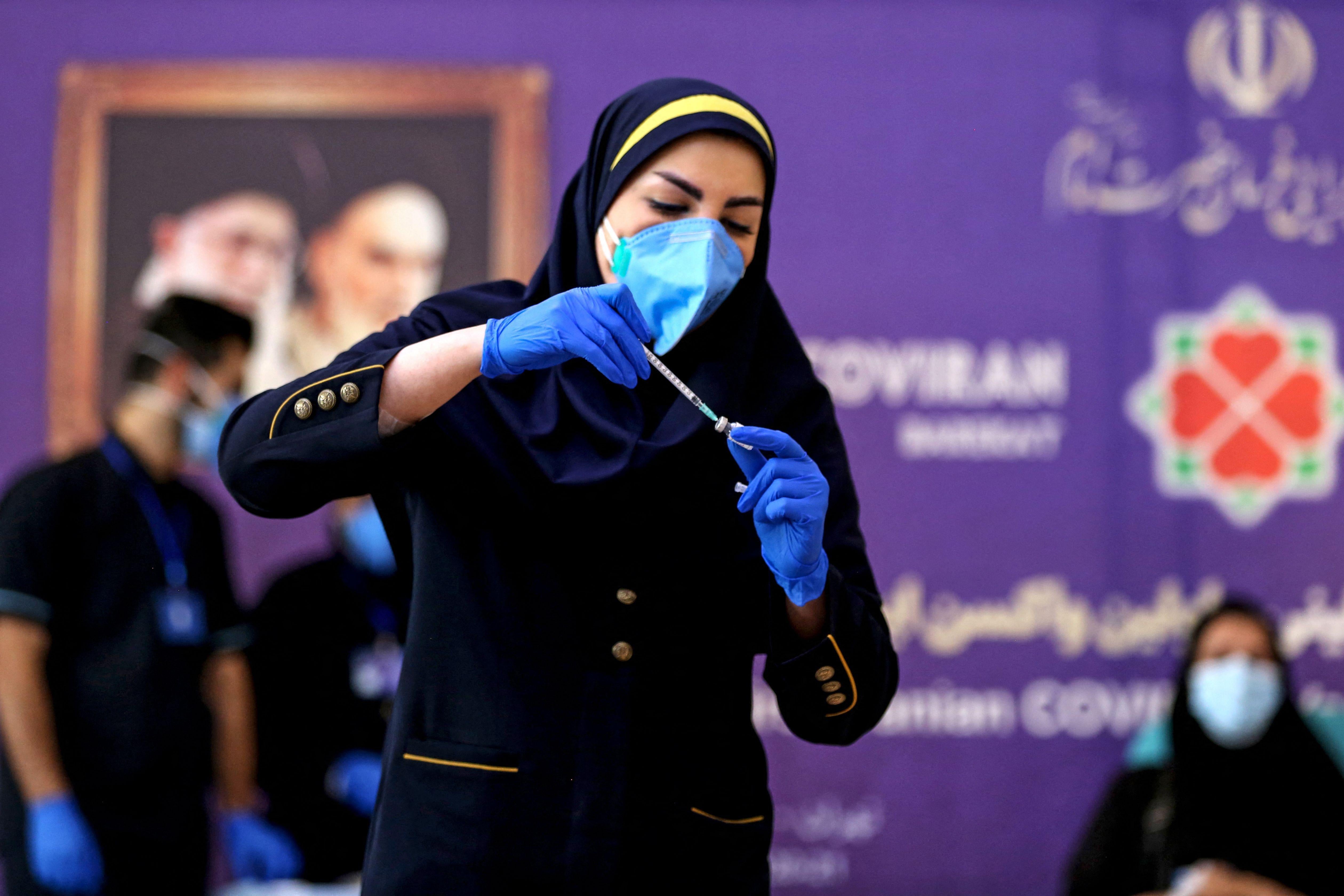 A woman wearing a headscarf, a mask, and gloves puts a syringe in a vial.