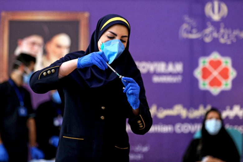 A woman wearing a headscarf, a mask, and gloves puts a syringe in a vial.