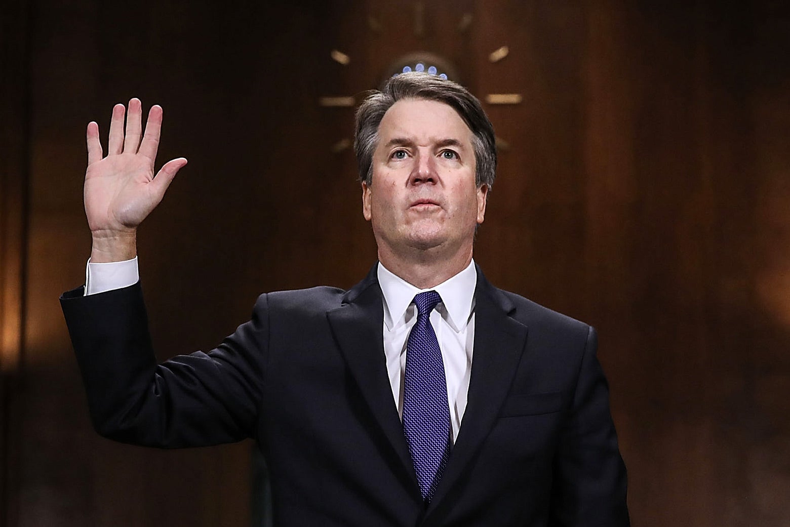 Brett Kavanaugh, wearing a dark suit and purple tie, holds up his right hand and looks forward.