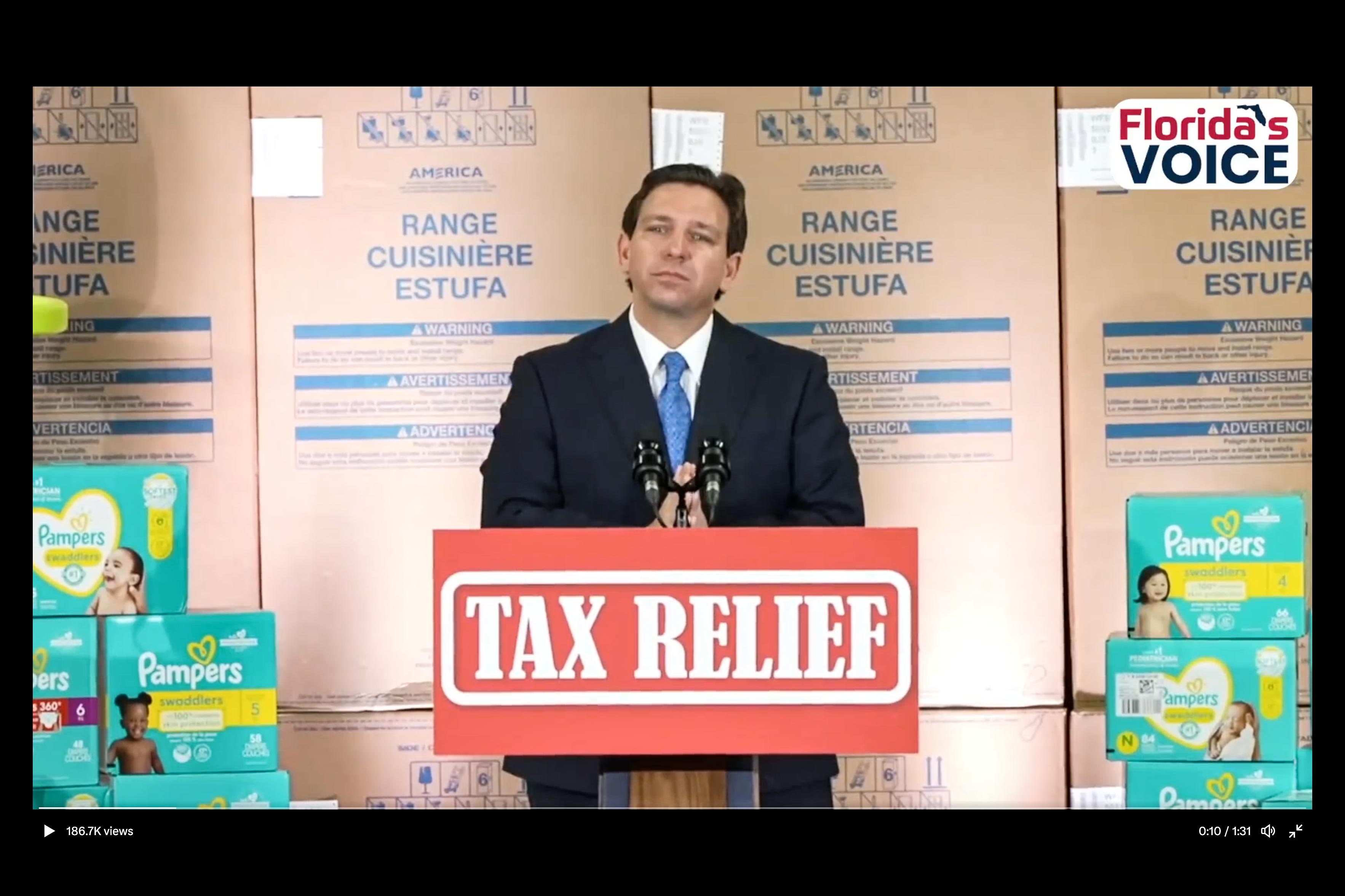Ron DeSantis, wearing a suit, speaks in front of a backdrop of cardboard boxes containing gas stoves. On each side of him are piles of Pampers diaper boxes.