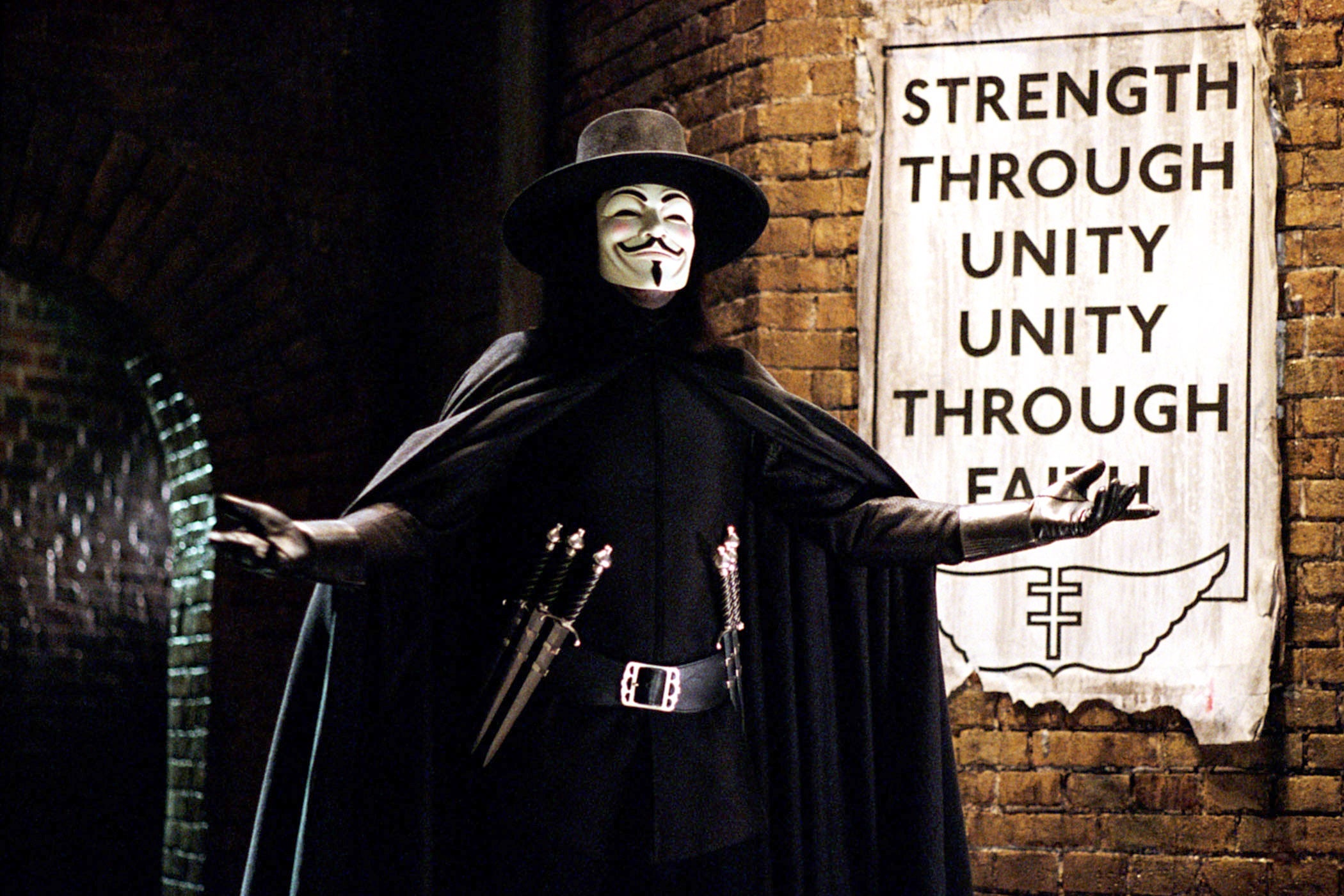 Hugo Weaving stands with his arms outstretched in his cape, top hat, tool belt with daggers, and Guy Fawkes mask, in front of a sign that says "Strength Through Unity, Unity Through Faith." 