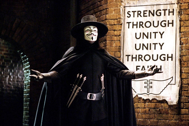 Hugo Weaving stands with his arms outstretched in his cape, top hat, tool belt with daggers, and Guy Fawkes mask, in front of a sign that says "Strength Through Unity, Unity Through Faith." 