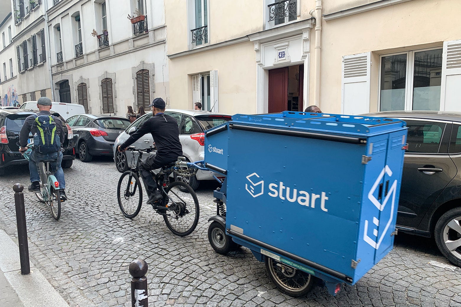 A delivery worker for Paris delivery company Stuart bikes down a cobblestone street with a small blue trailer in tow.