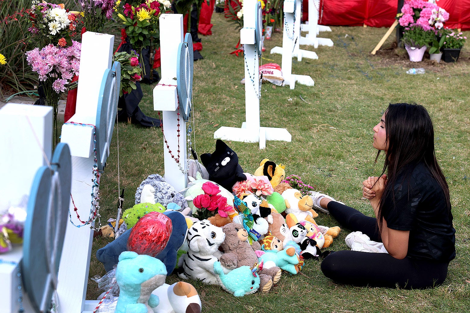 A young woman sits on the ground and prays in front of a row of white crosses with a pile of stuffed animals left in front of it.