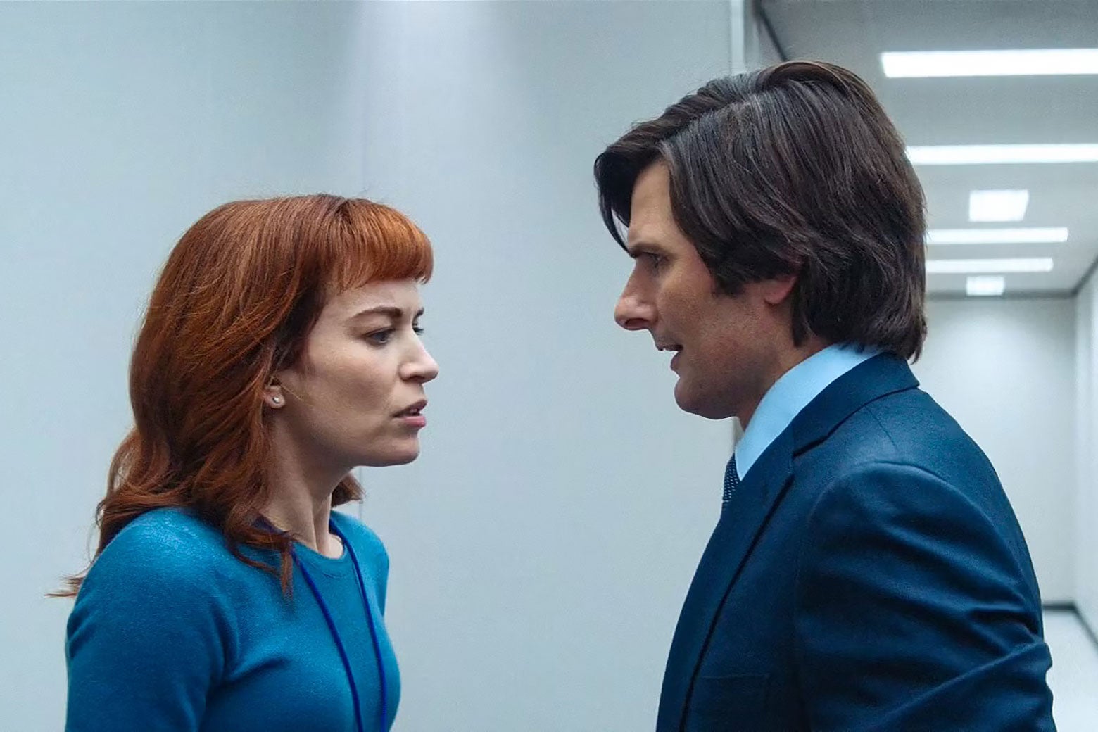 A woman and a man huddle in a workplace hallway and have a covert, urgent conversation.