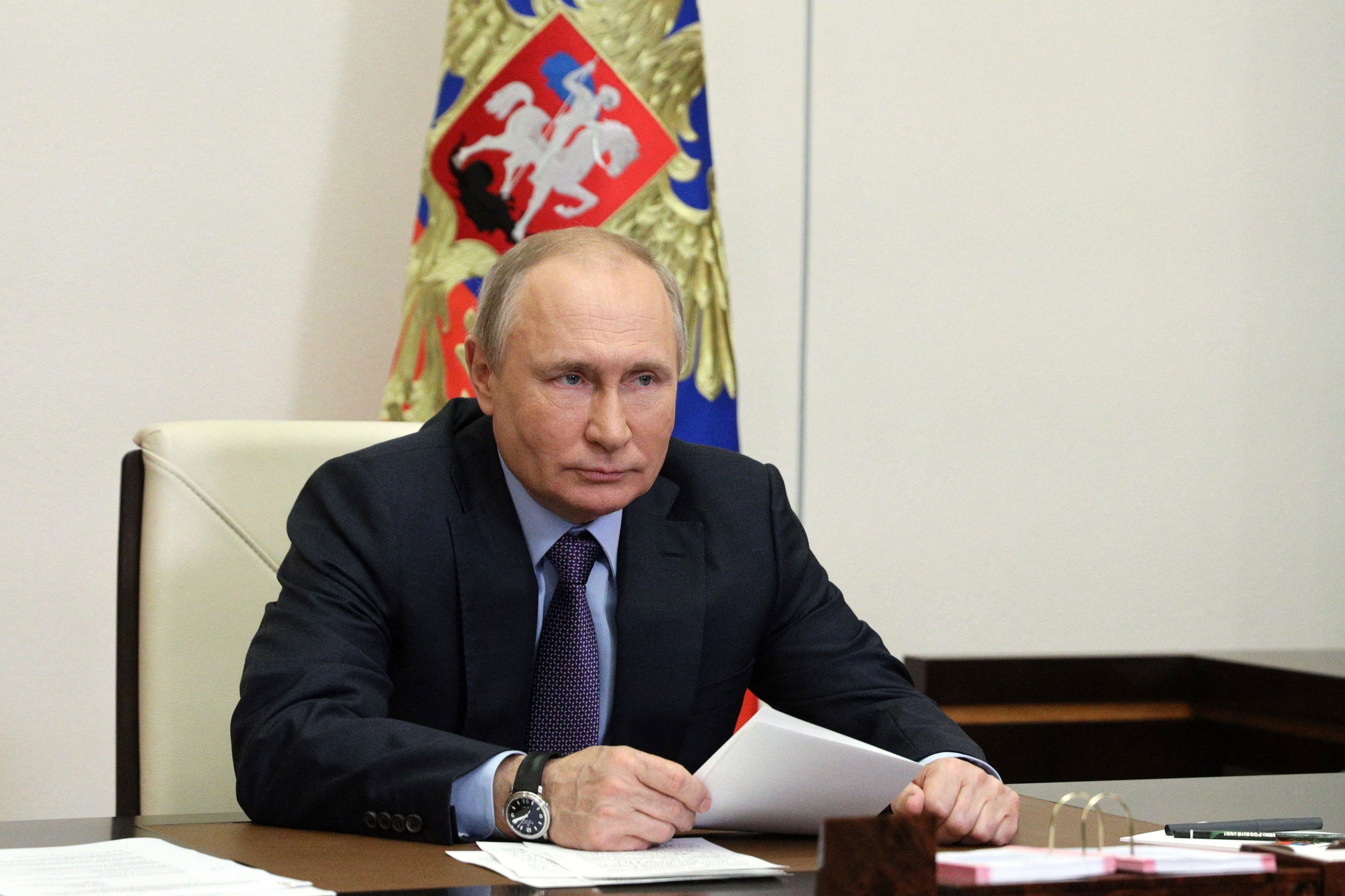 Vladimir Putin sits at a table holding a piece of paper, with a flag behind him.