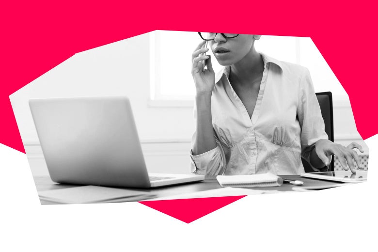 A woman wearing glasses sits at her desk and looks at her computer while holding her phone up to her ear.