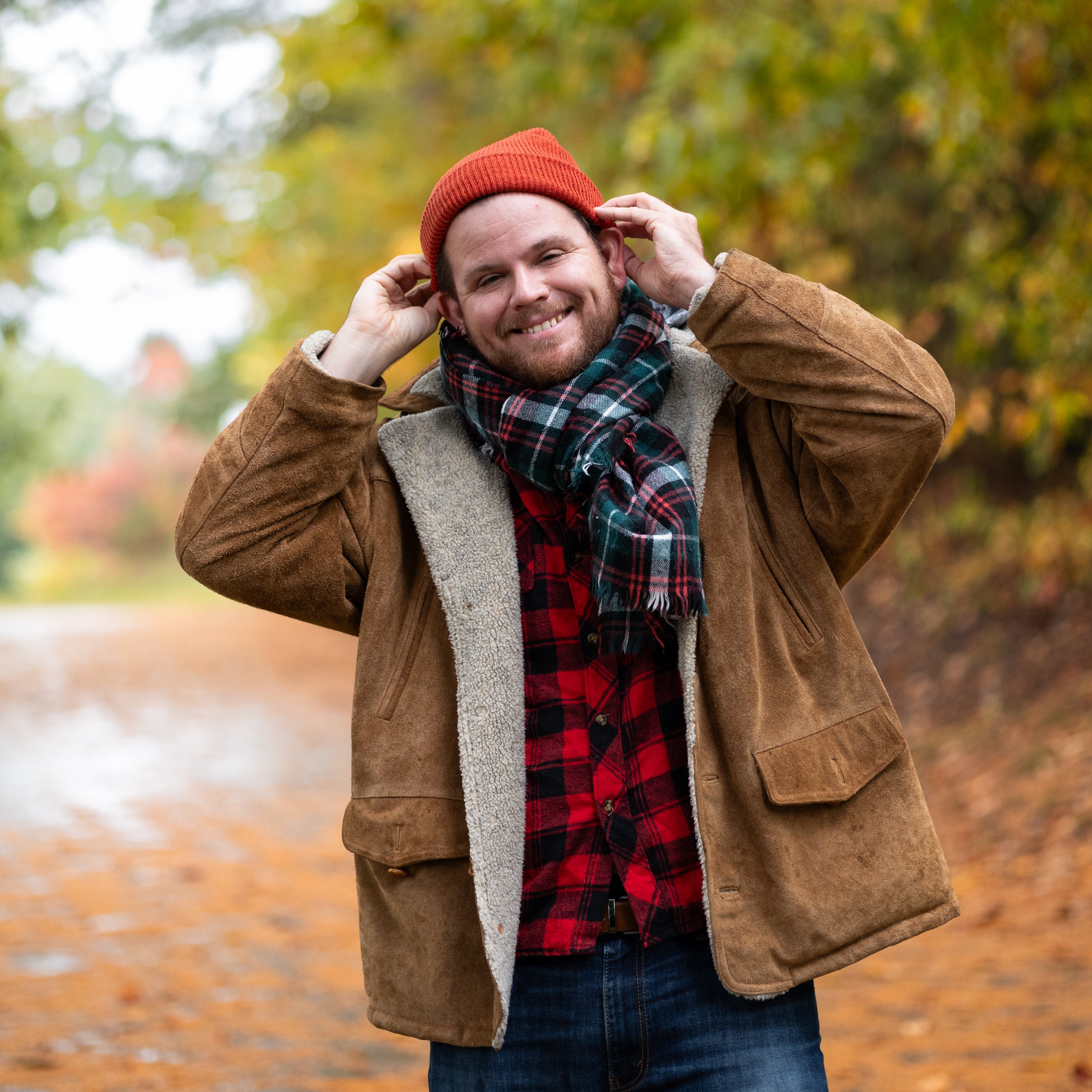 Luke pulls a hat over his head while smiling at the camera. He wears a tartan scarf and brown jacket over a checkered red-and-black shirt.