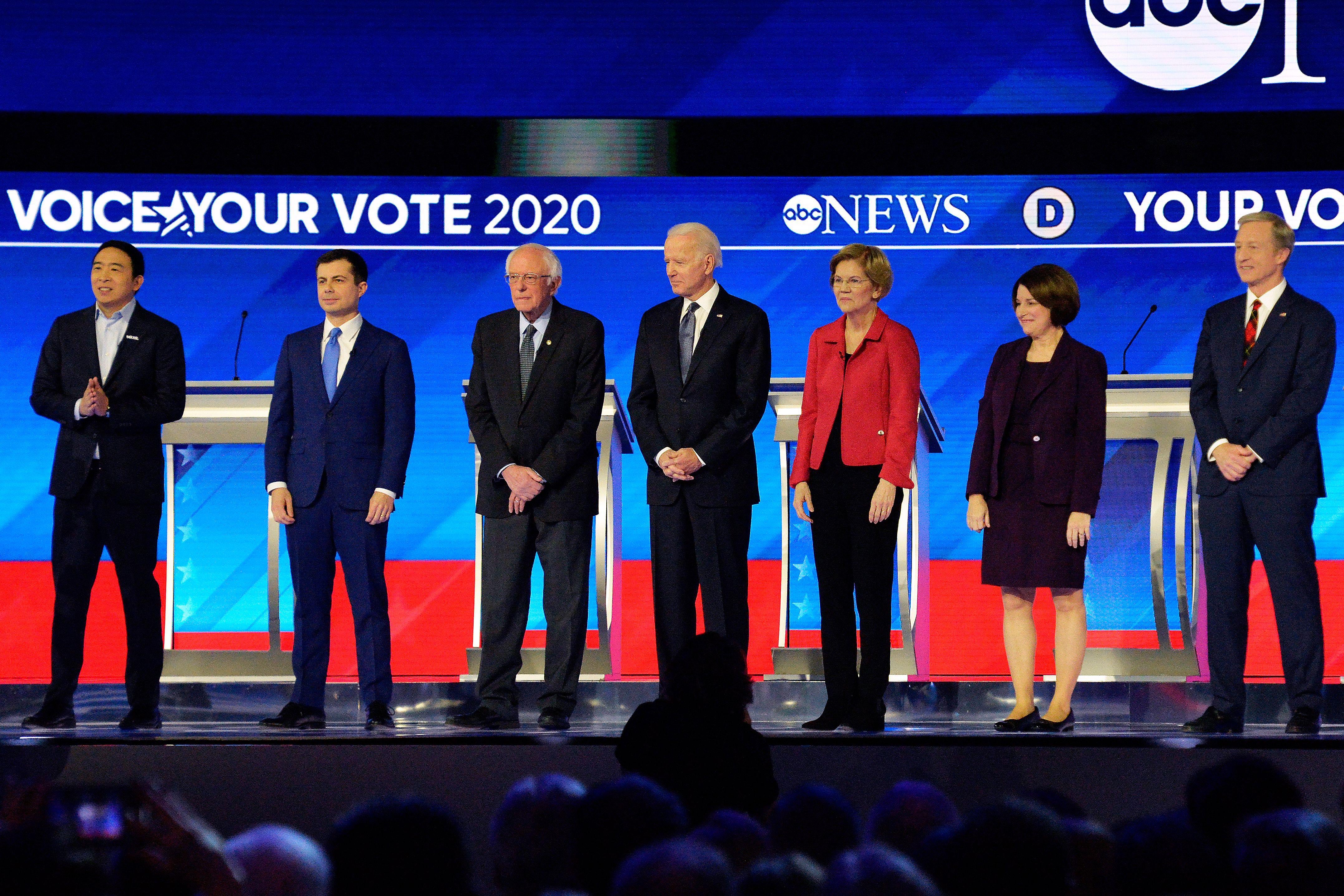 Andrew Yang, Pete Buttigieg, Bernie Sanders, Joe Biden, Elizabeth Warren, Amy Klobuchar, and Tom Steyer all stand on a stage.