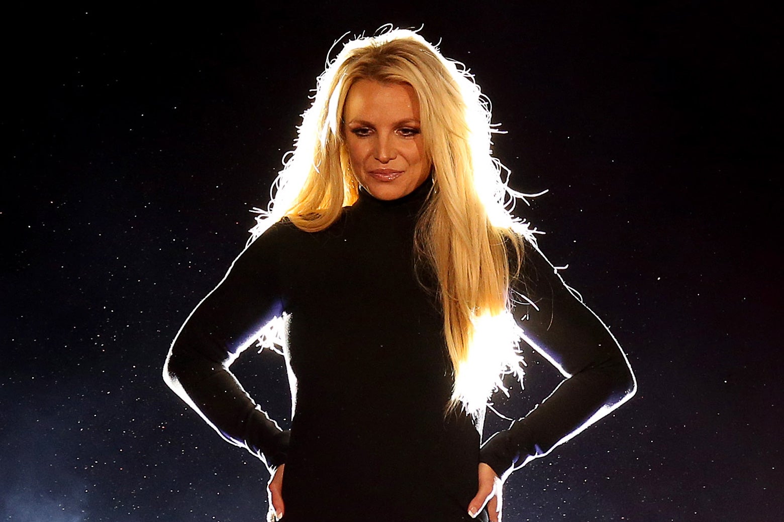 Britney Spears looking pensive onstage in a black dress with her hands on her hips facing the audience