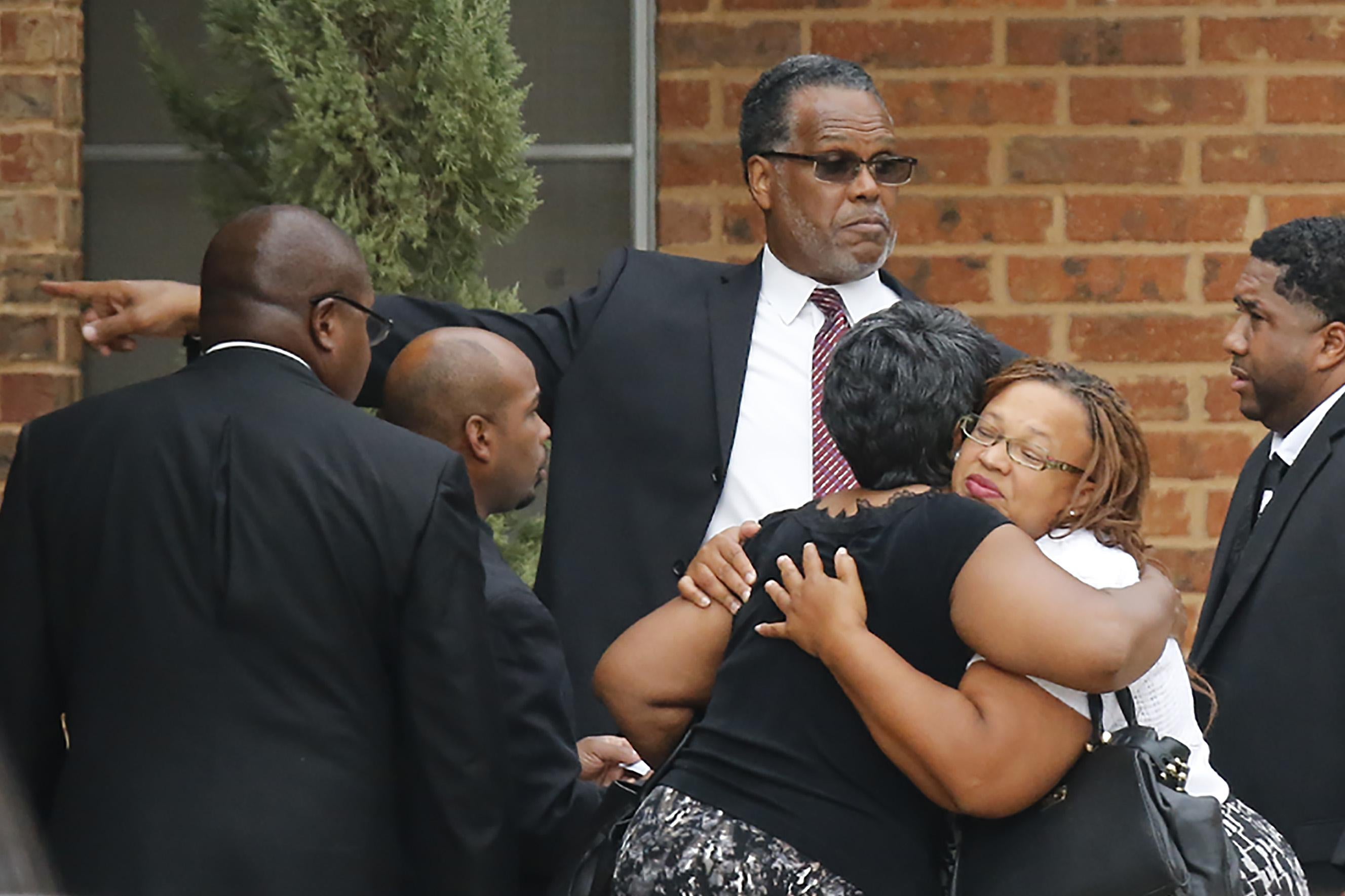 Two women, dressed in funeral clothes, embrace. A man in a suit directs people toward the service.