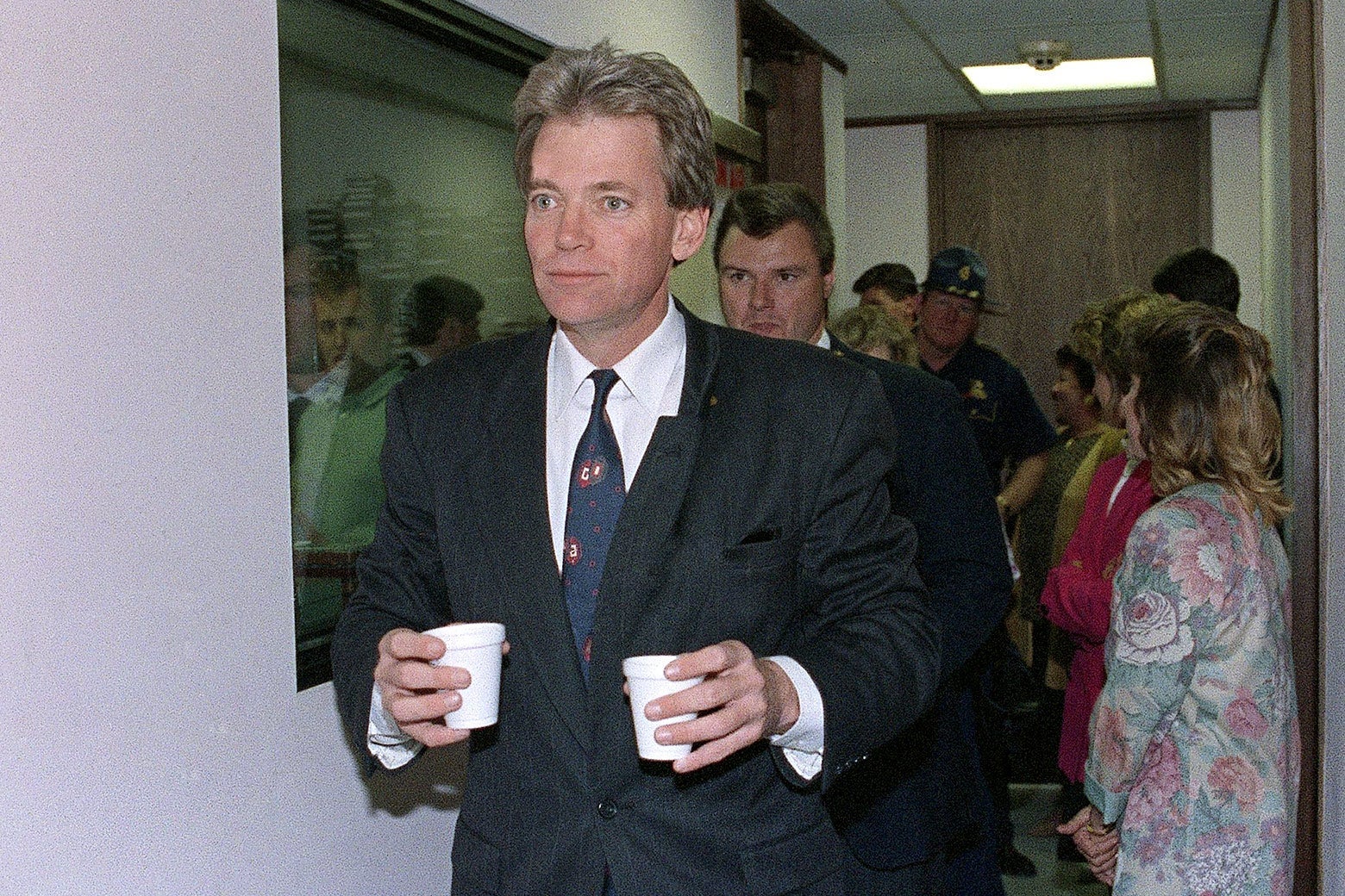 David Duke carrying two little styrofoam cups. A radio studio is seen, along with people milling about, behind him.