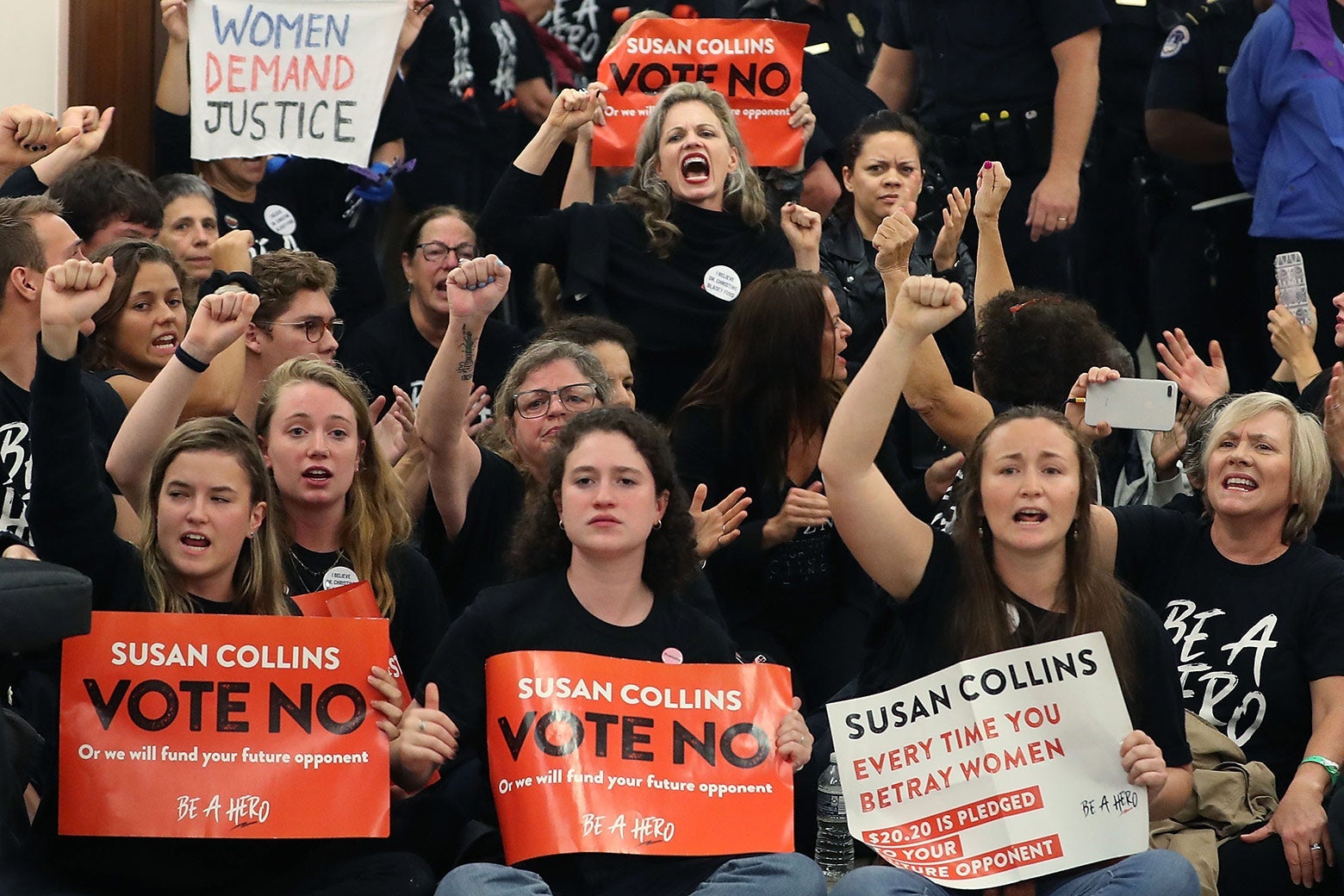 A crowd of women hold up their fists and hold signs that say, "Susan Collins Vote No."