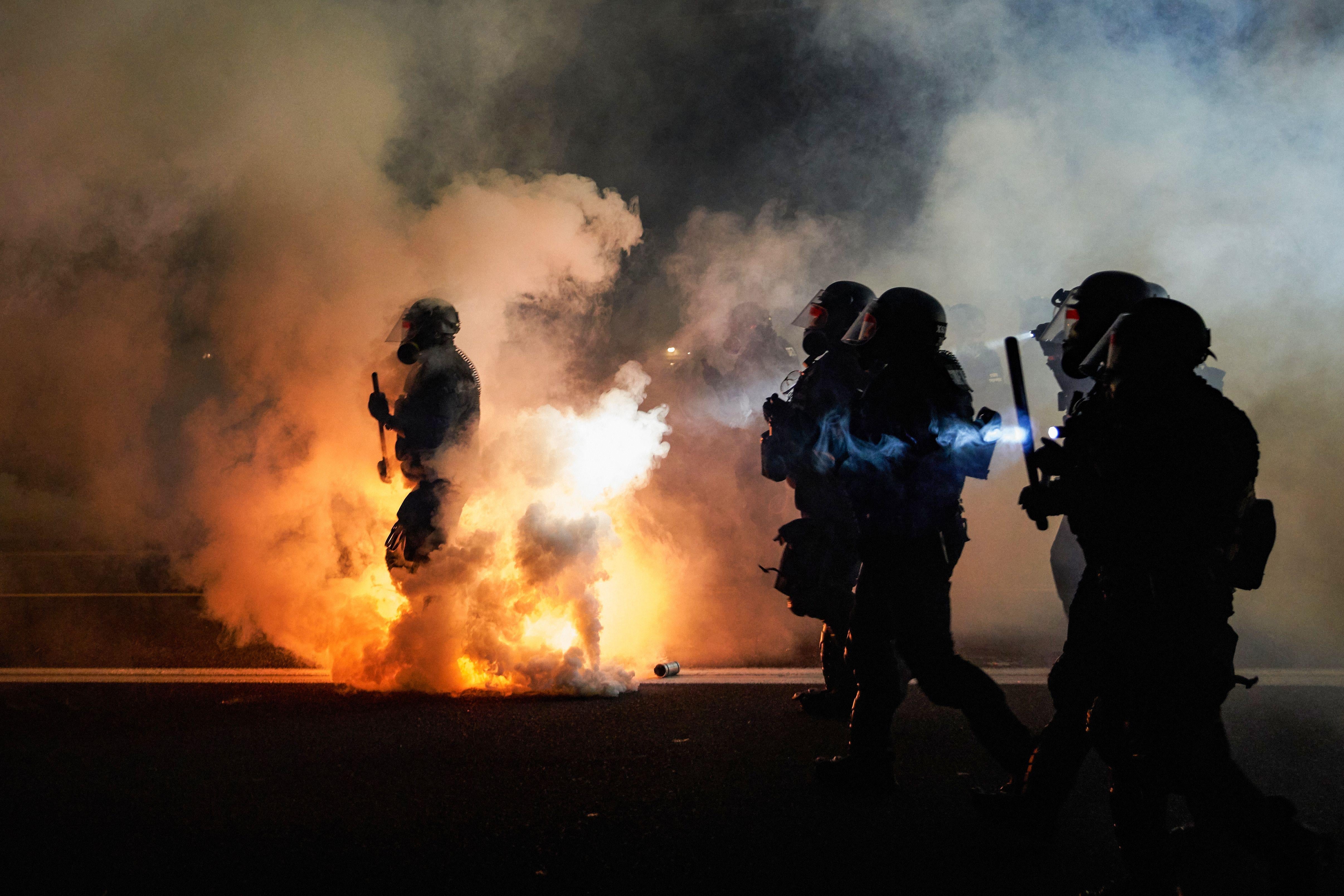 Police wearing riot gear and holding batons walk through tear gas at night