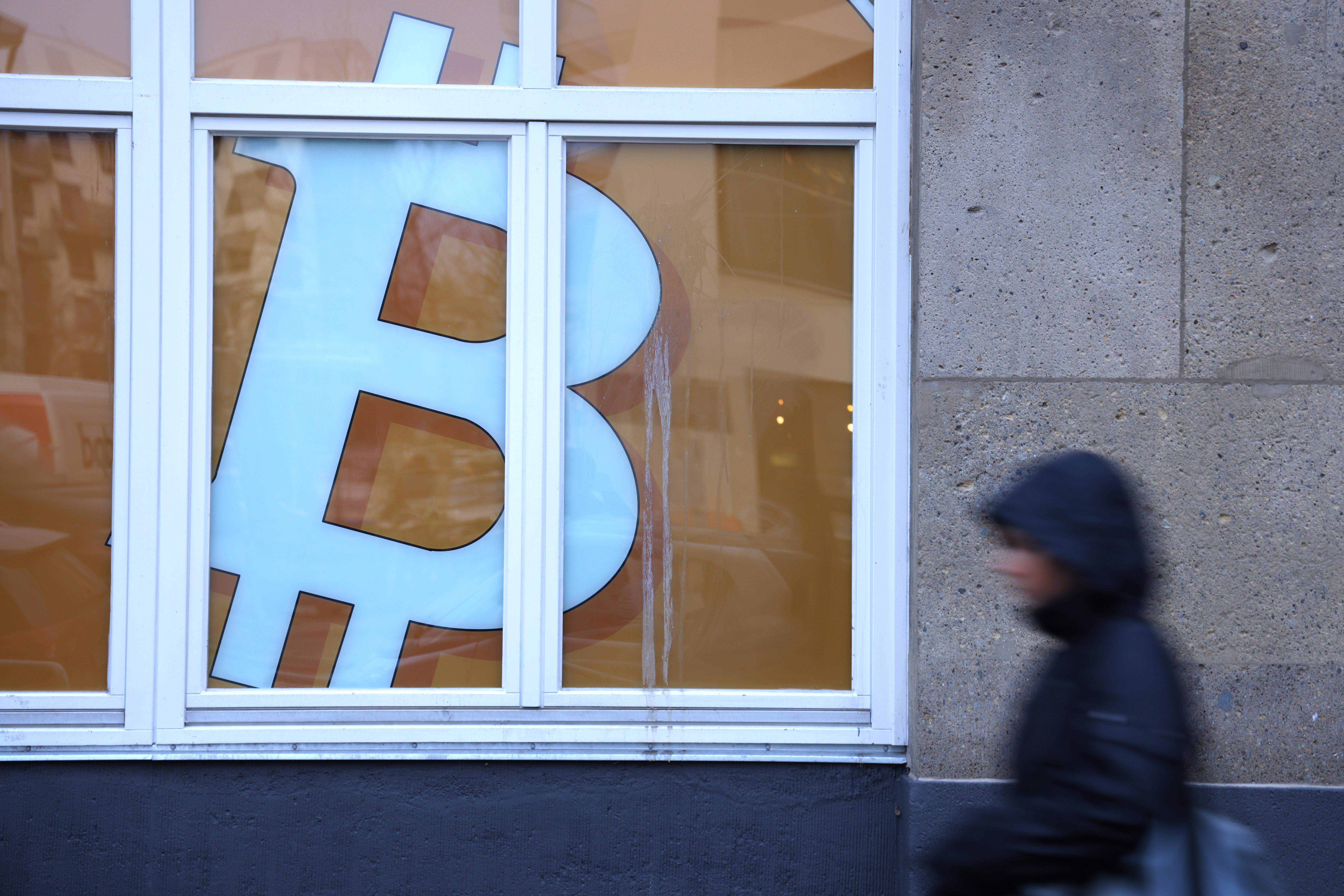 Person in a puffy coat walks past a Bitcoin symbol in the window of a building