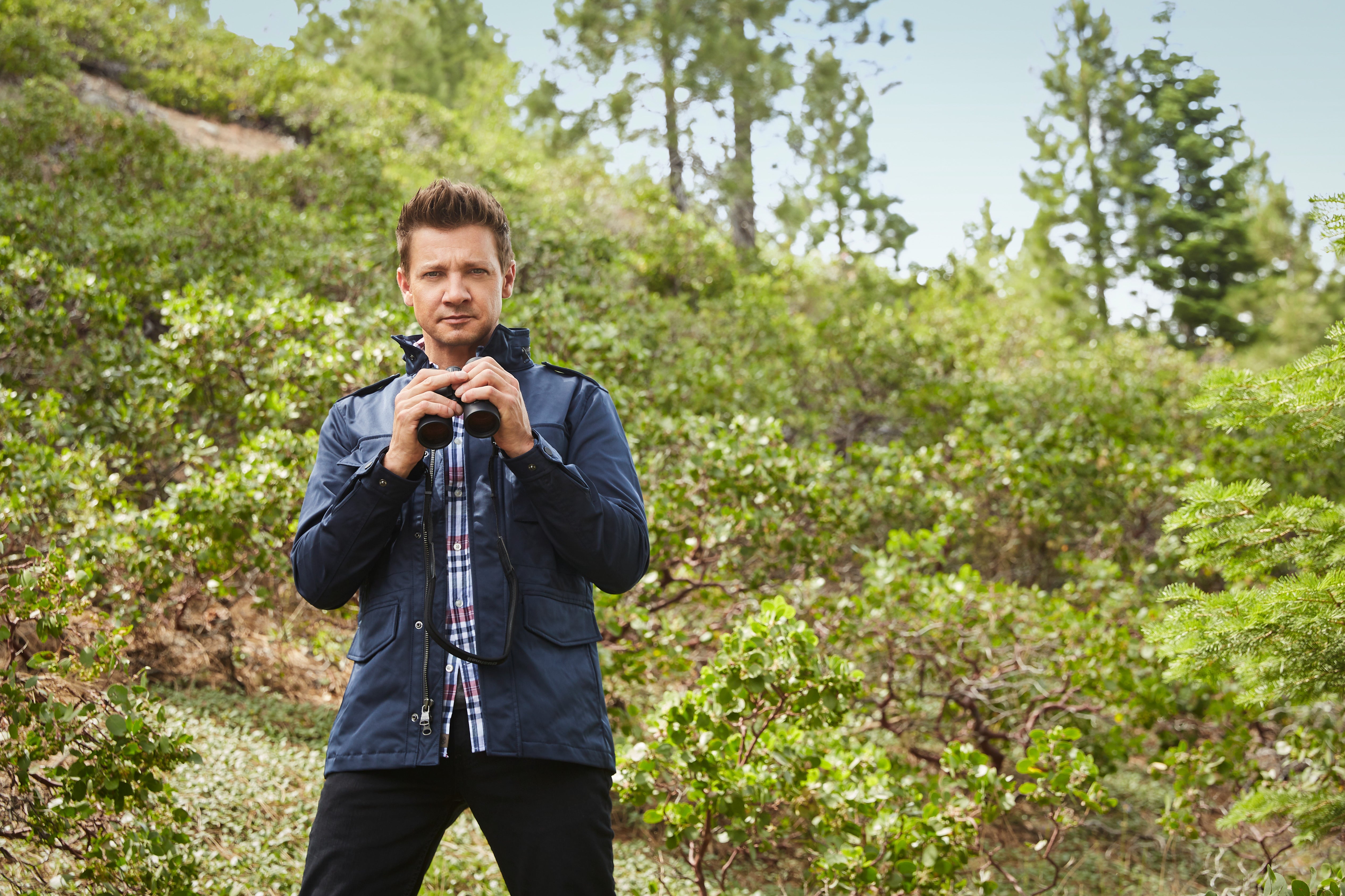 Jeremy Renner stands holding binoculars but again not looking through them.