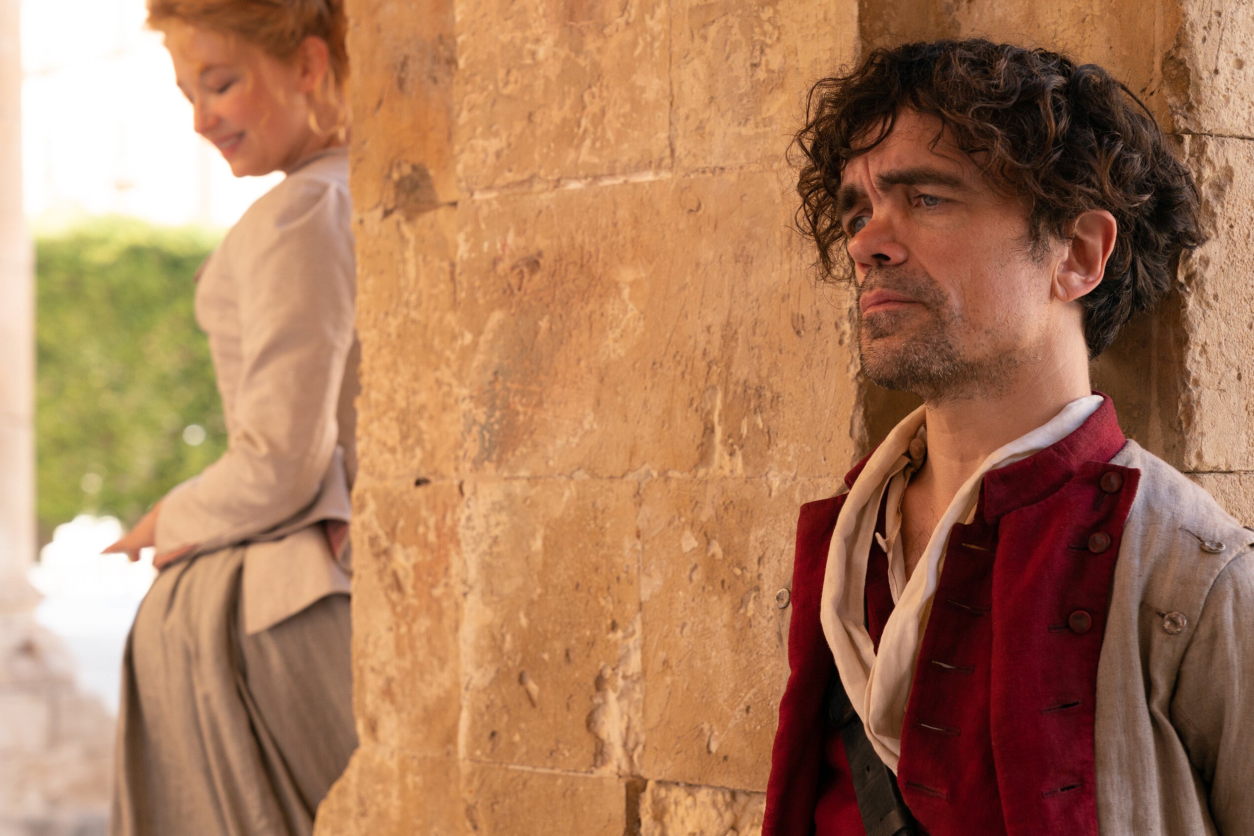 On the right of the frame: Peter Dinklage stands behind a stone pillar in period-piece clothing, looking straight ahead with a sad and pained look on his face. On the left, a woman stands on the other side of the pillar, unable to see Peter Dinklage. She smiles slightly, looking down. 