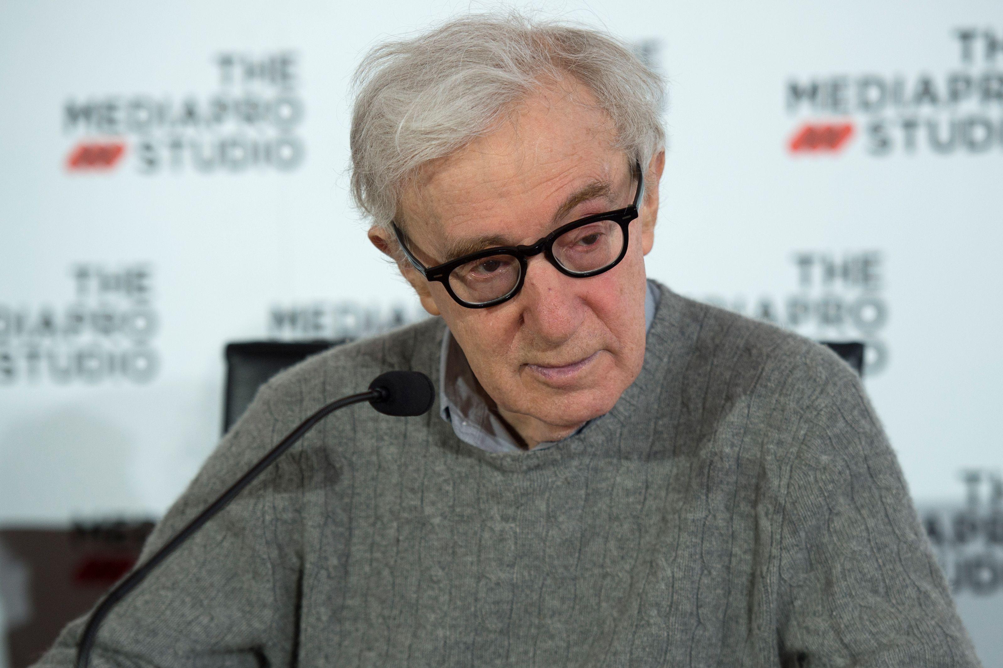 Woody Allen sits behind a microphone, wearing a gray sweater with a collared shirt under it.