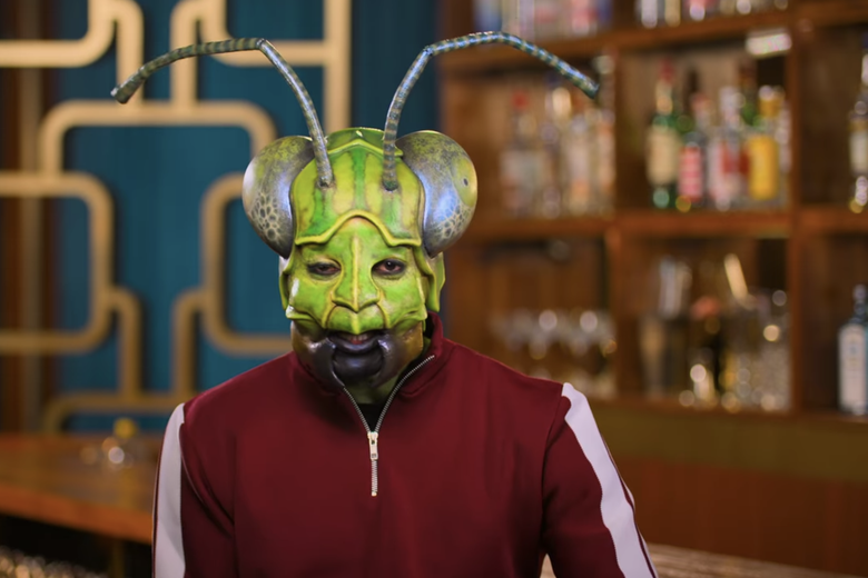 Man in red jacket wearing realistic praying mantis mask.