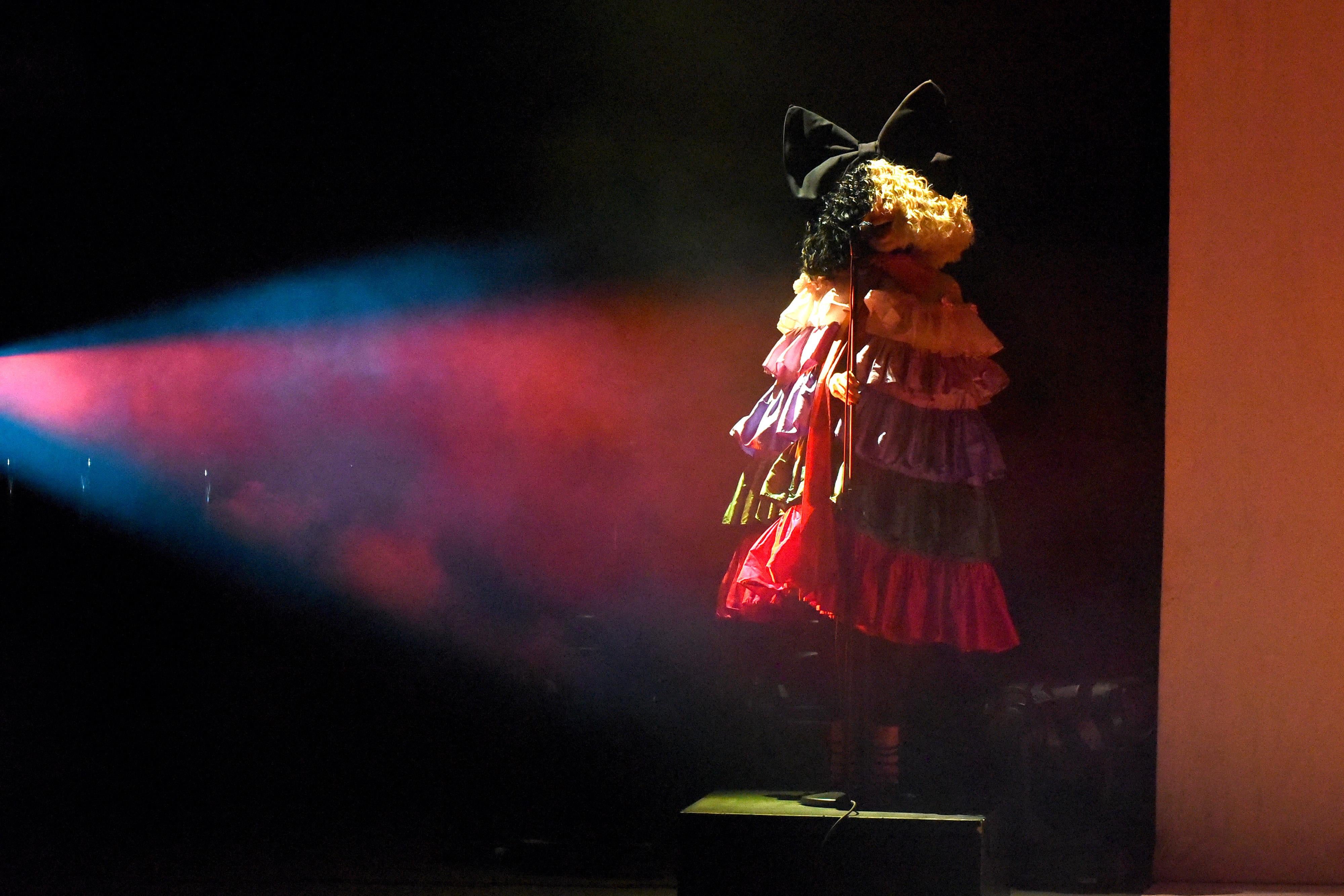 Sia, wearing a multilayered ruffled dress and a large black-and-white wig that covers her entire face, stands alone on a stage.