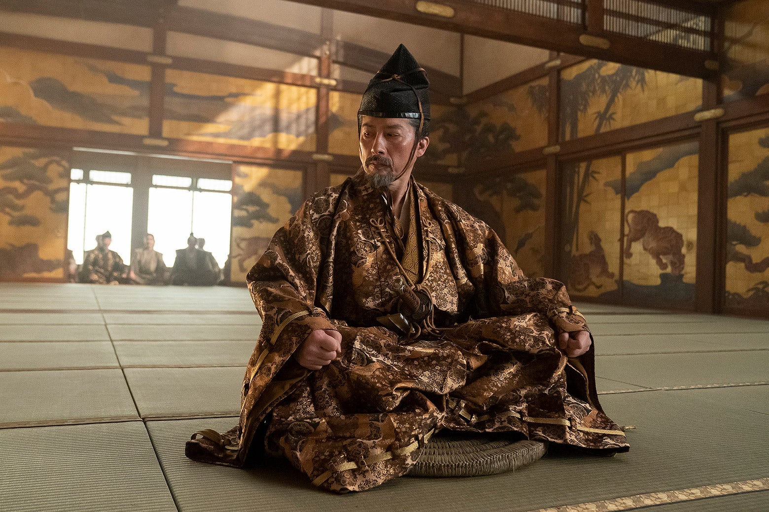 A gentleman in a silk robe sitting legs crossed in the middle of a dojo.