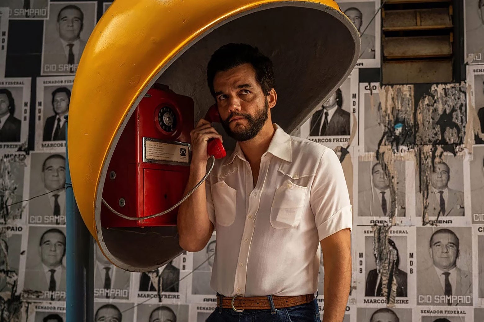 A young man in a white button-down shirt places a call from a mustard-yellow, 1970s-era pay phone.