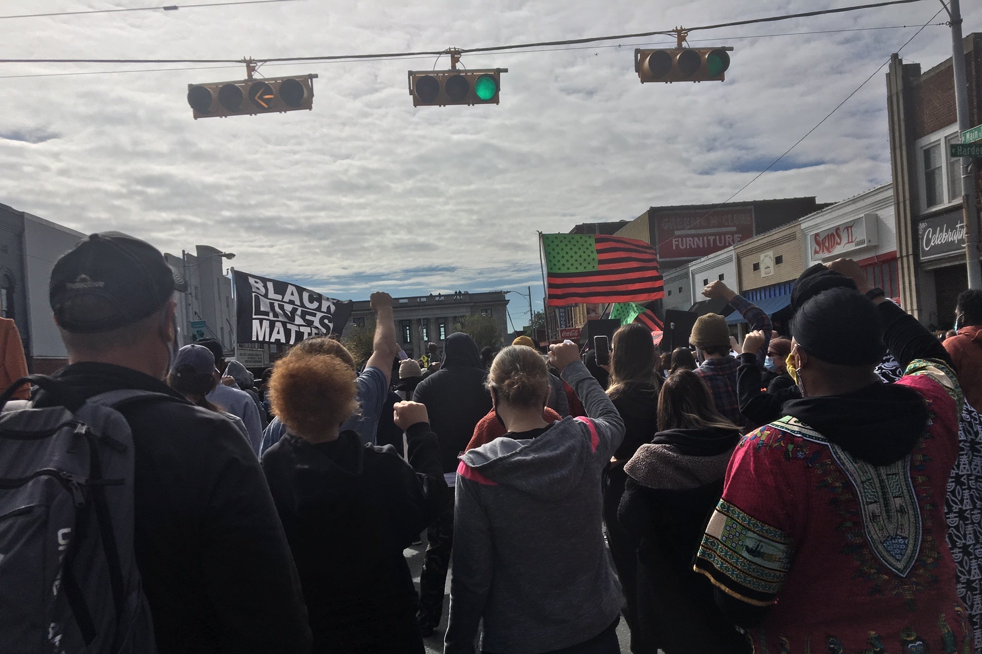 A crowd of people march down a street with a red, green, and black American flag and a Black Lives Matter flag
