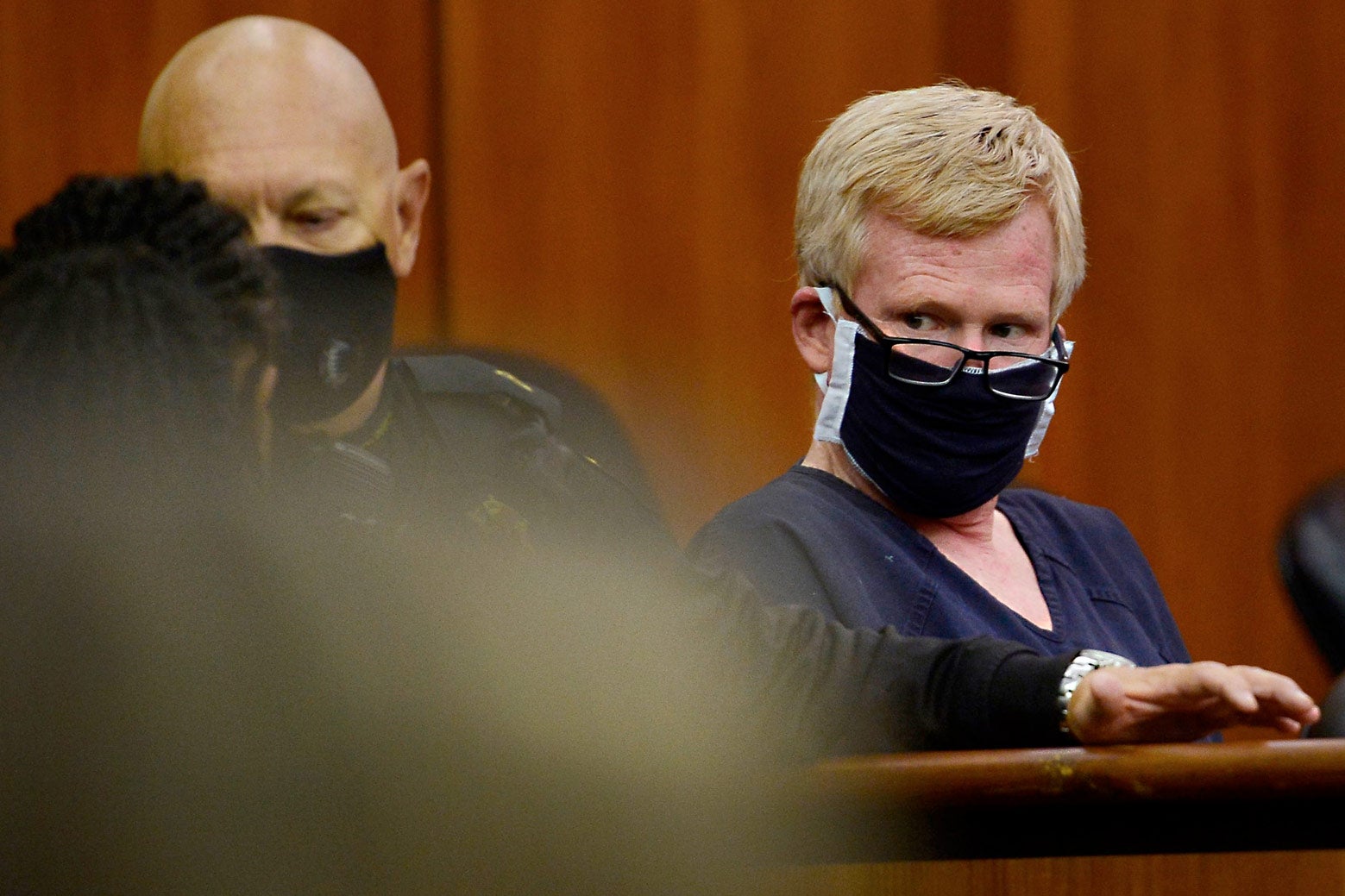 The red-haired Murdaugh, wearing a blue prison jumpsuit and glasses over a black COVID mask, looks to his right while seated in a courtroom.