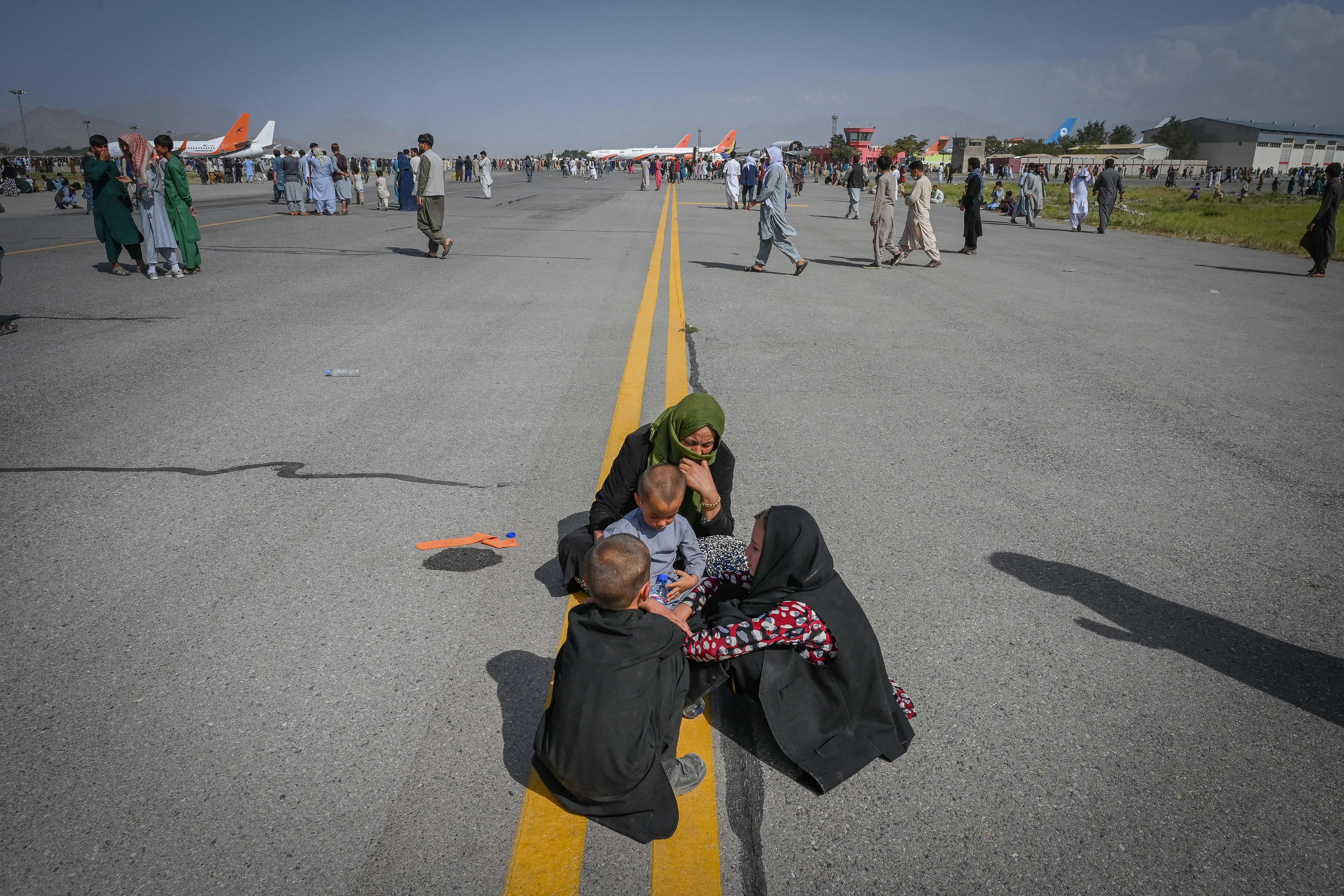 Afghans stand outside on the runway. In the foreground a family sits on the tarmac.