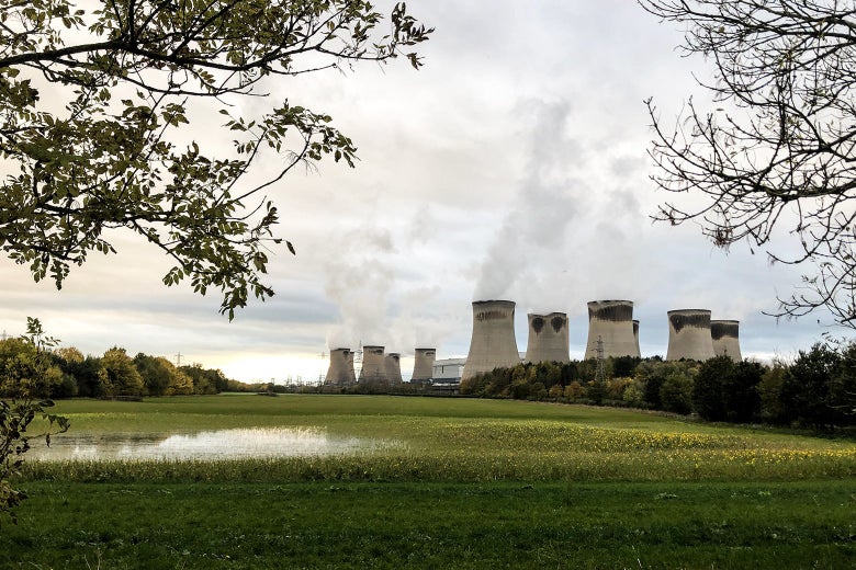 A smoking energy plant is seen behind a large meadow and clumps of trees.