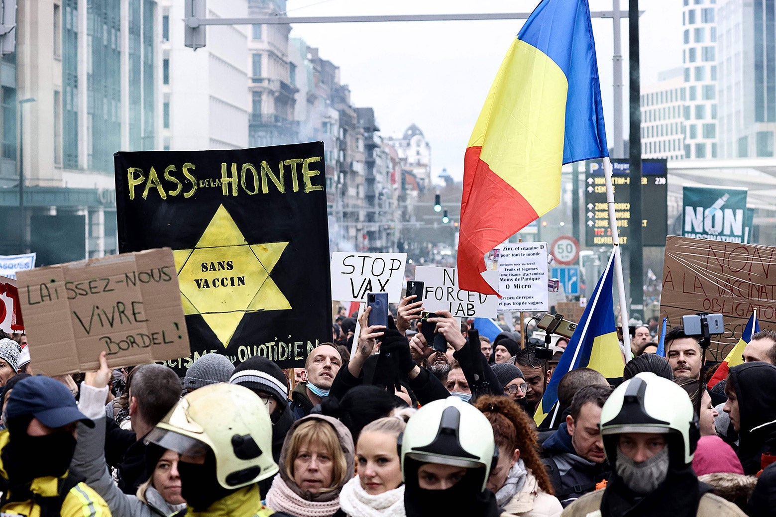 People march in the street holding flags and anti-vax signs including one with a yellow star