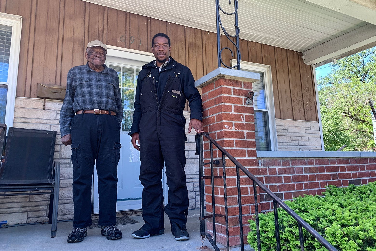 An elderly Black man in a plaid shirt and baseball cap and a younger Black man in Black overalls stand on a brick porch in front of a house.