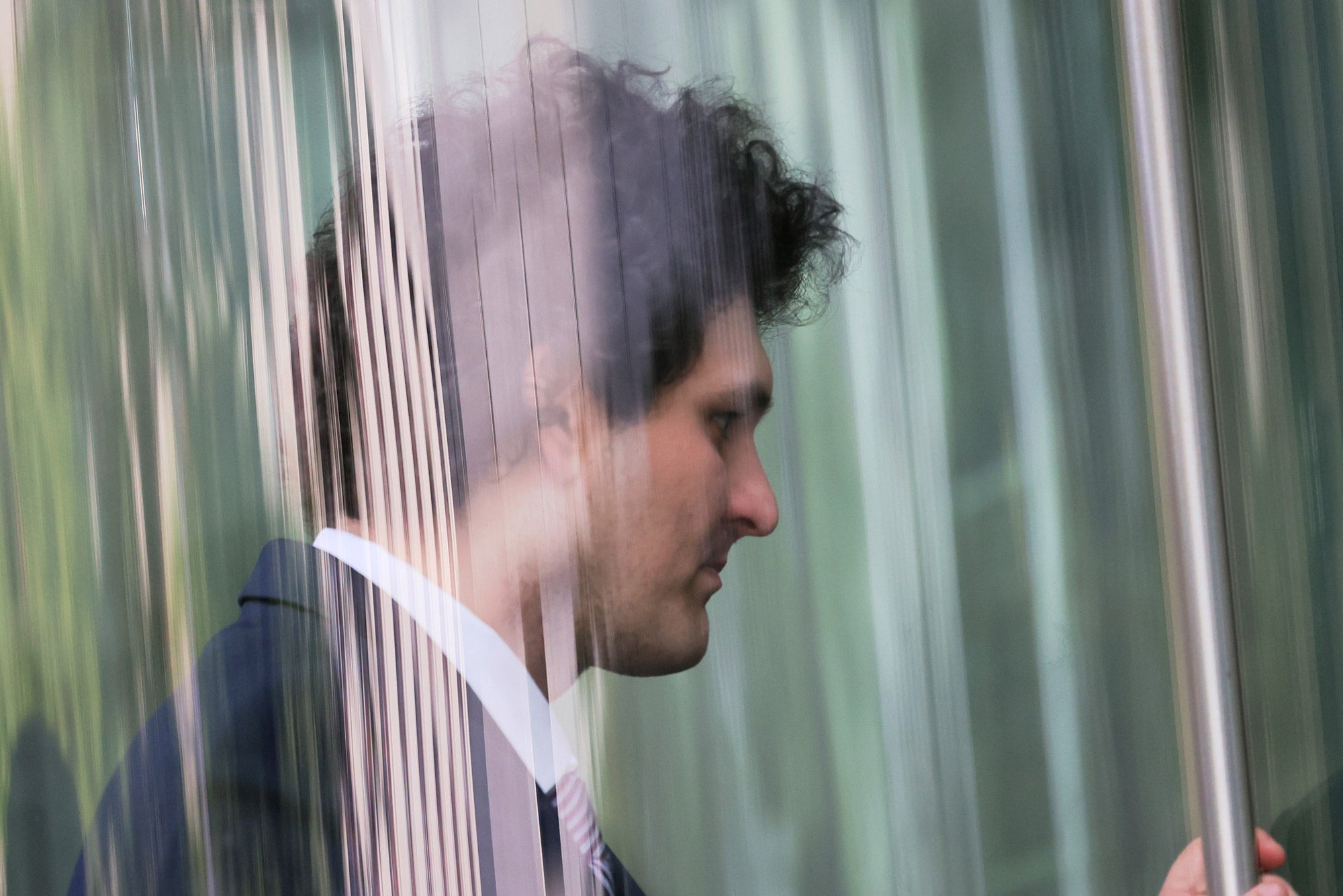 Sam Bankman-Fried, viewed through a pane of glass, enters a revolving door. He looks solemnly ahead.