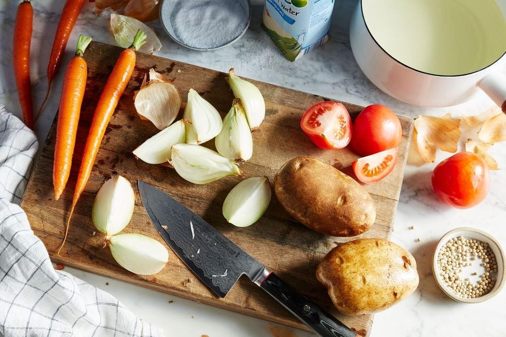 Potatoes, carrots, tomatoes, onions, and a knife on a wooden cutting board, surrounded by more veggies, dishes of salt and seeds, a pot, and a box of coconut water, all on a marble countertop