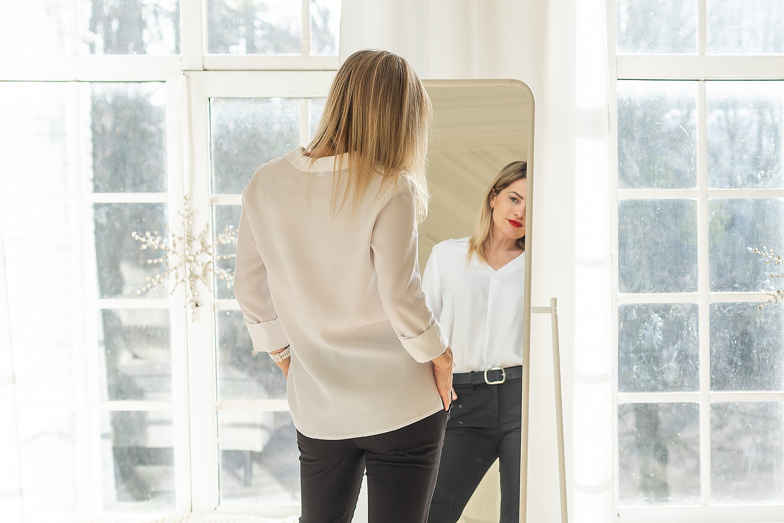 A blond woman looking in a full length mirror in a large room with natural light.