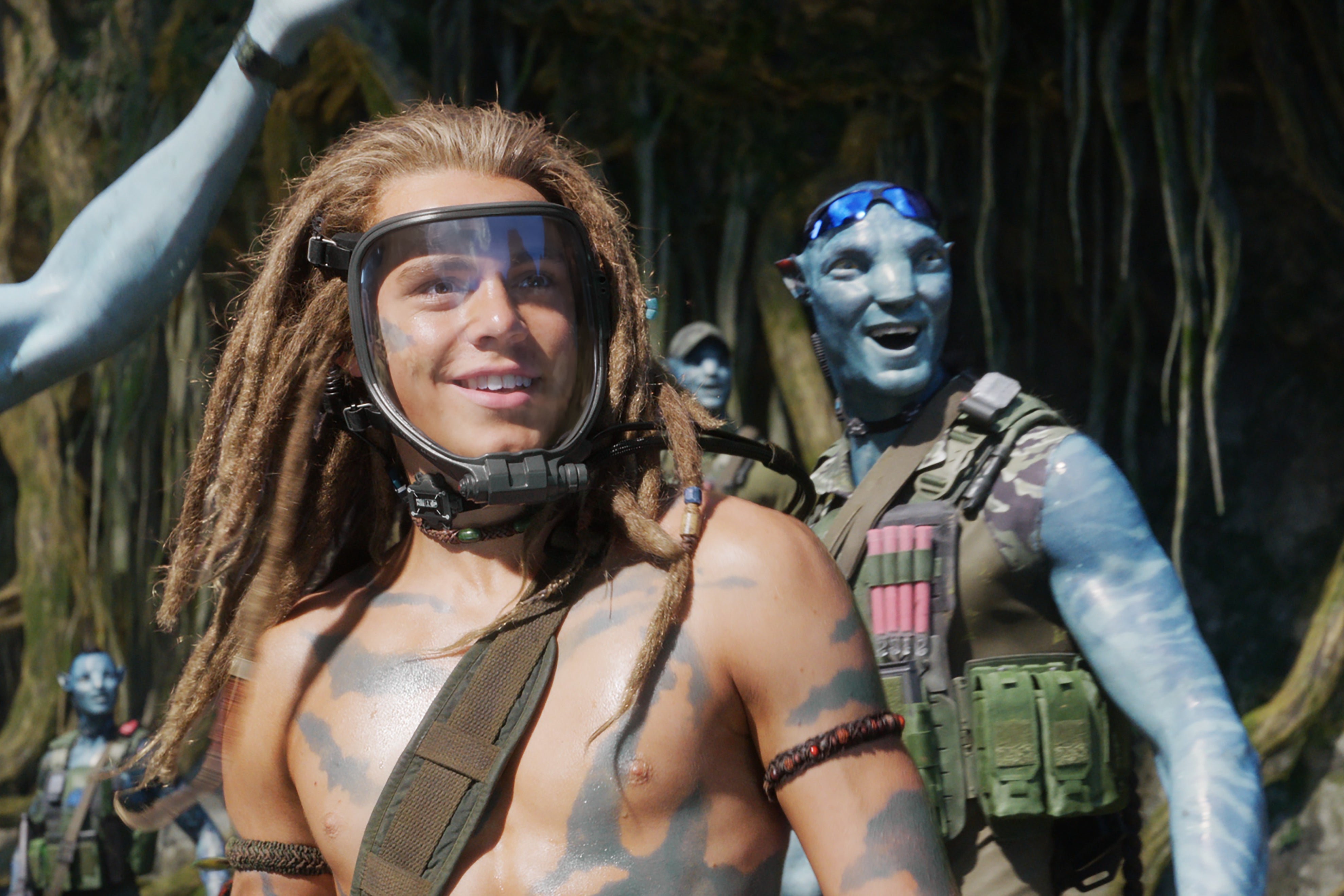 A shirtless, tattooed white guy with beads around his arms, blond dreads on his head, and a face-covering gas mask, looks on in awe, as blue cat people in camo gasp and smile behind him