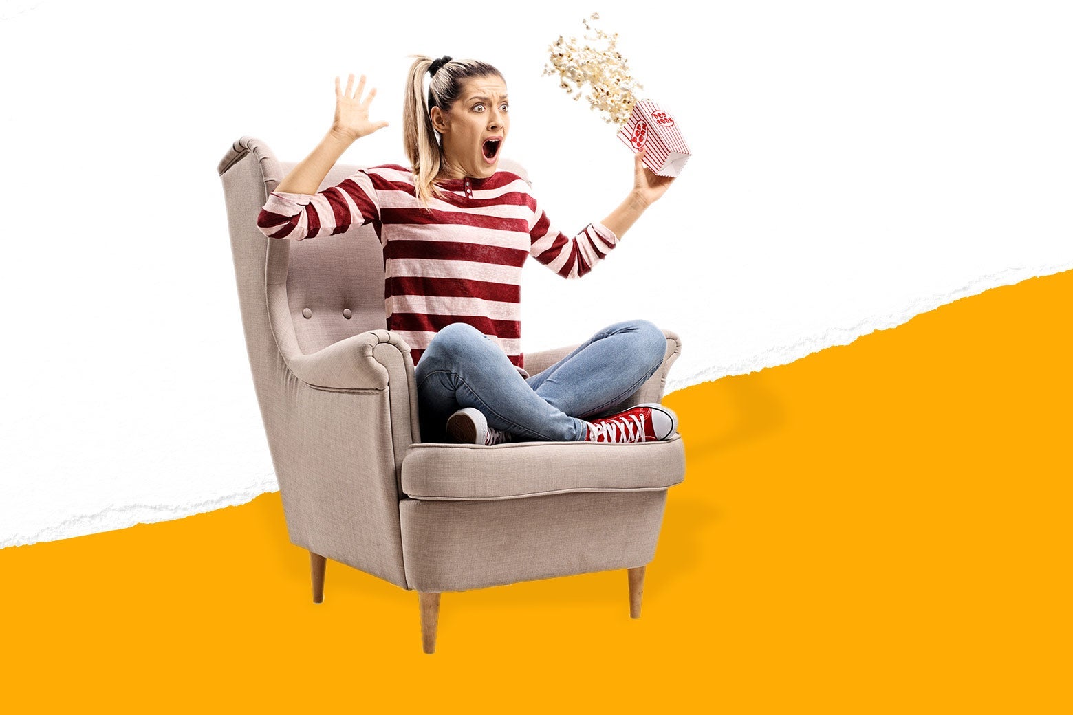 A woman sitting cross-legged on an easy chair throws her arms up, making the popcorn she was holding in her hand spill.